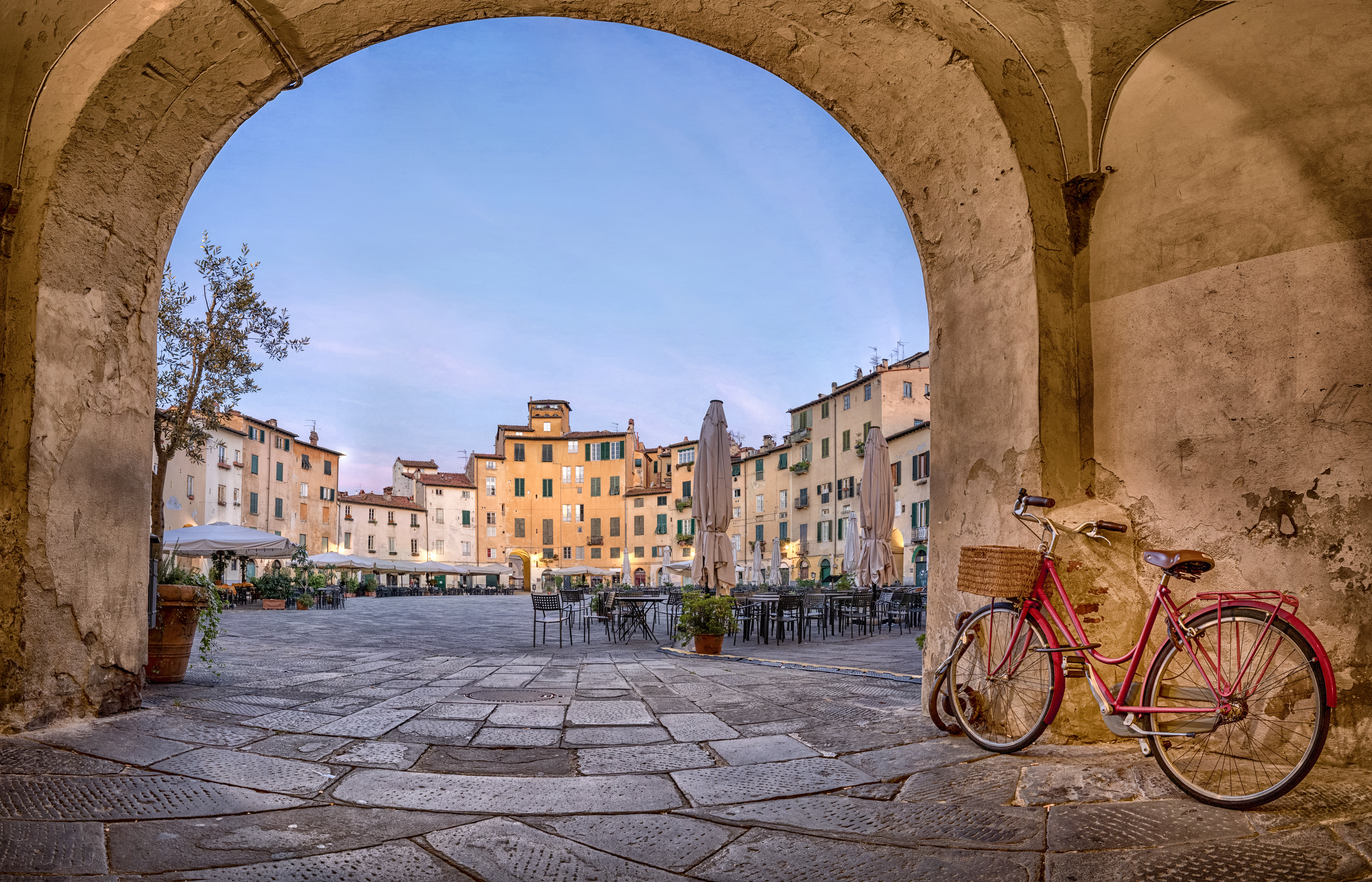 Magisk morgenstemning ved Piazza dell'Anfiteatro i Lucca, Toscana, set gennem gammel stenbue med rød cykel og historiske bygninger omkranset af caféer