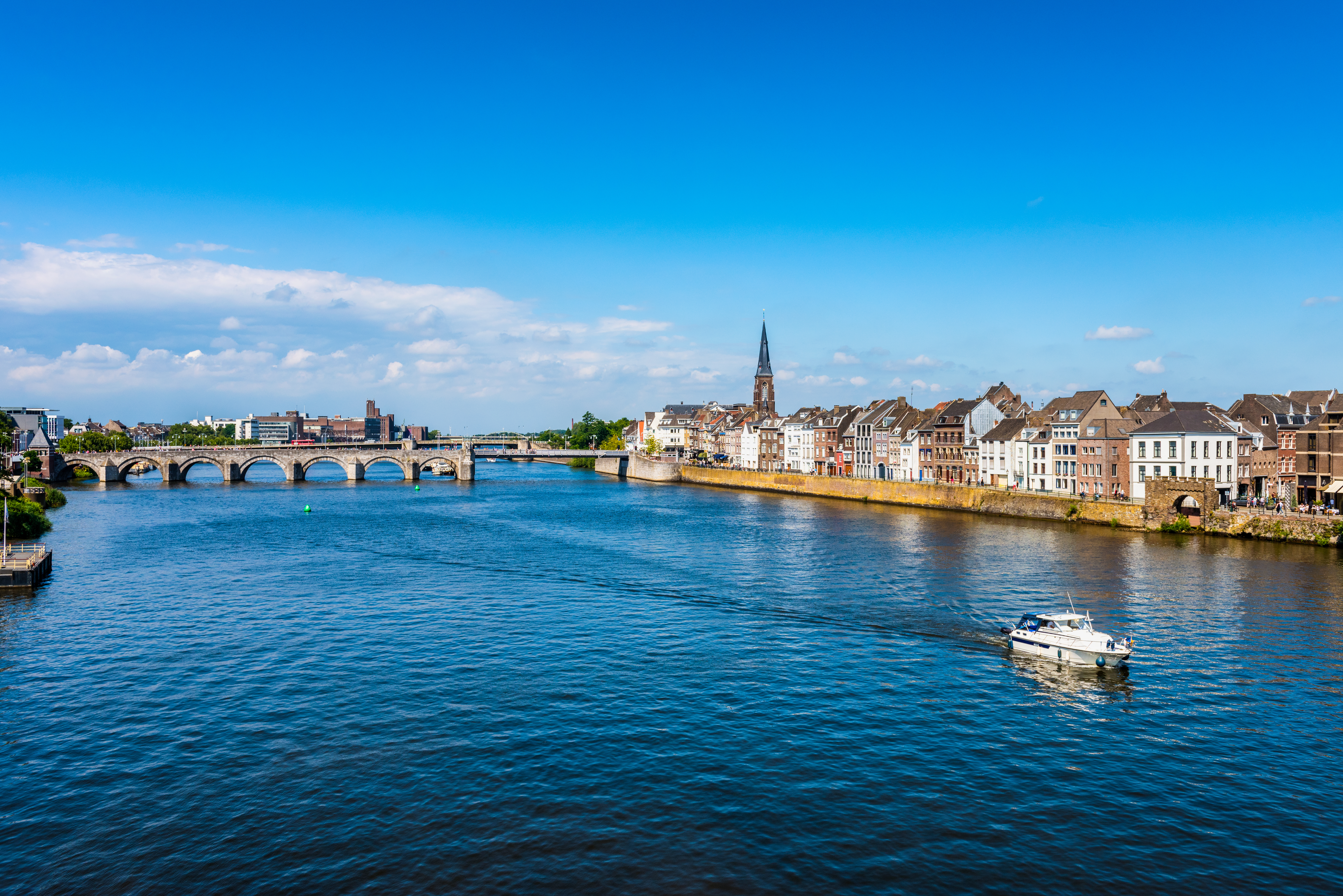 Idyllisk udsigt over Maastrichts historiske centrum langs Meuse-floden med motorbåd, middelalderlig stenbro og kirkespir i Limburg