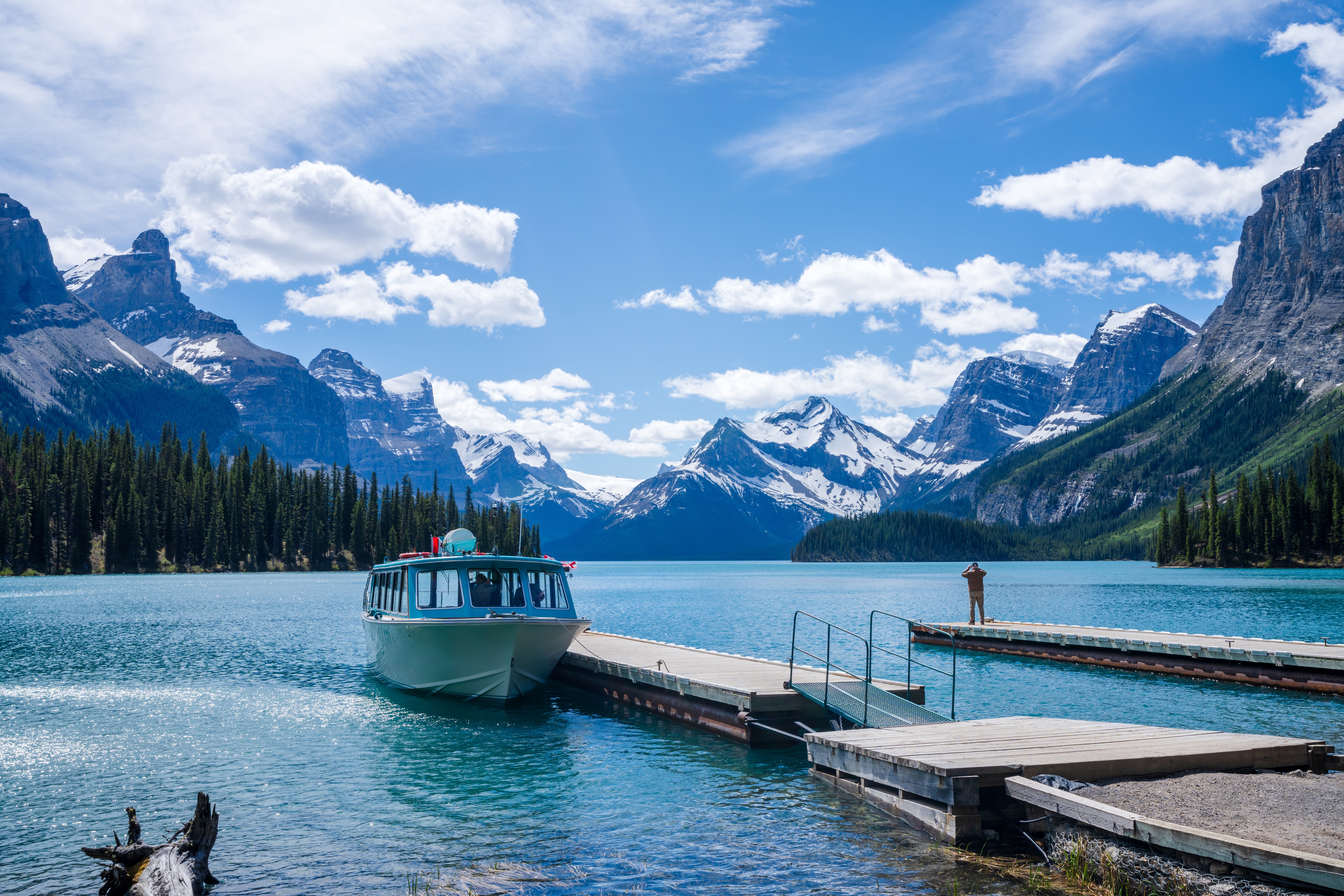 Turkisblå Maligne Lake med krydstogtbåd ved træbrygge og dramatiske snedækkede Rocky Mountain-toppe under blå himmel i Jasper National Park, Canada
