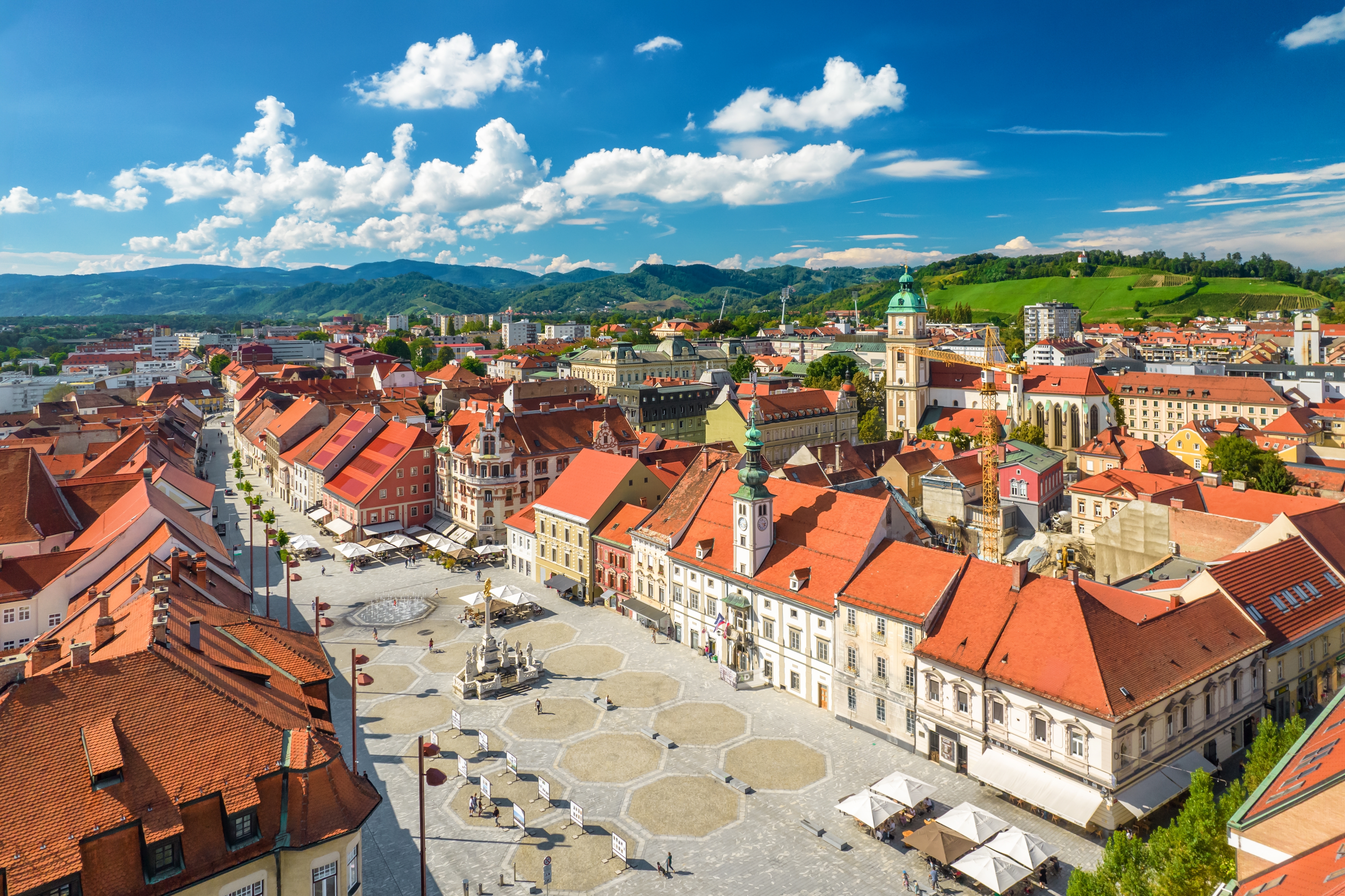 Smukt luftfoto af Maribors historiske bytorv i Slovenien med farverige bygninger, røde tage og den berømte pestsøjle på en solrig sommerdag