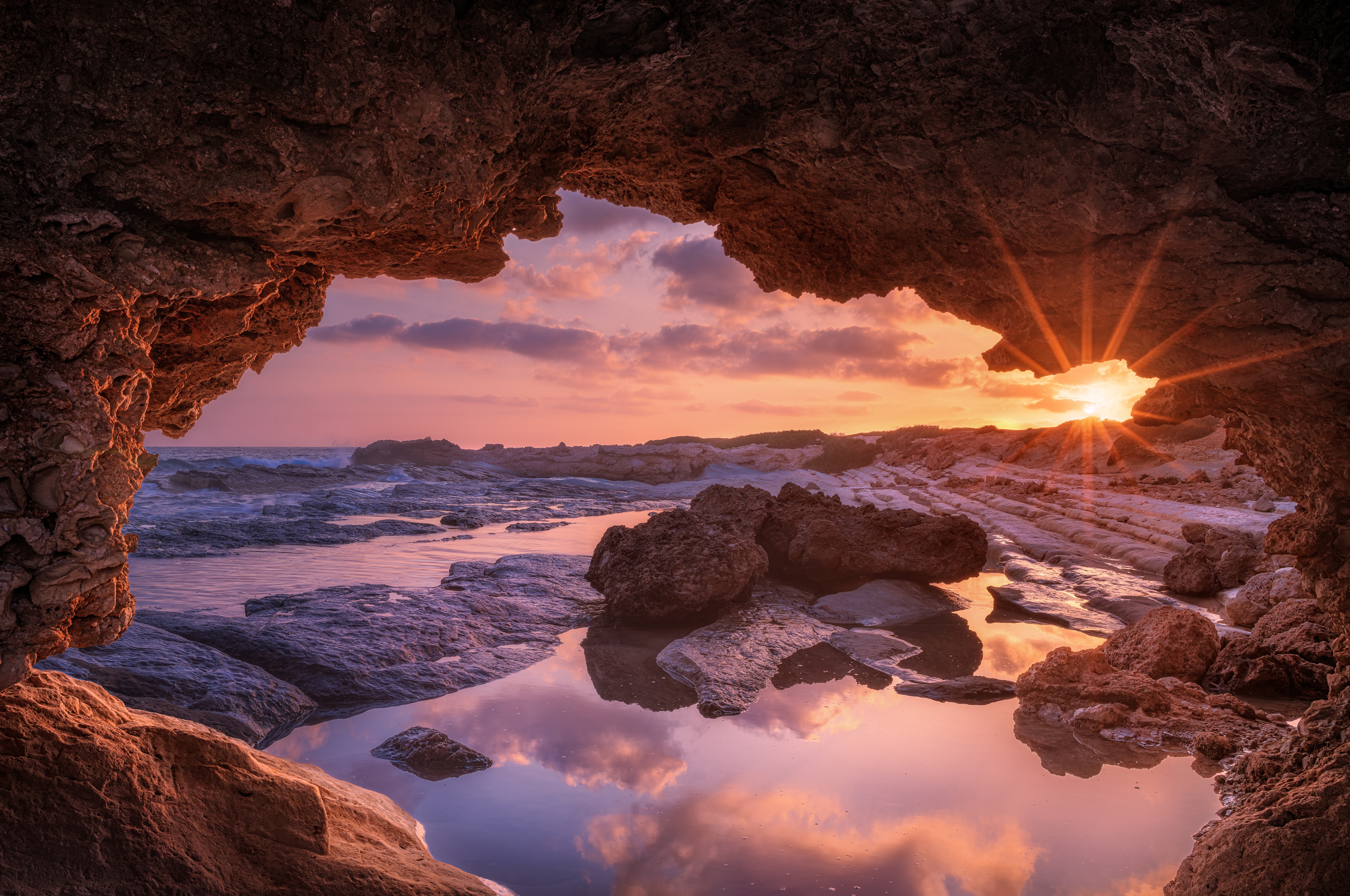Fantastisk udsigt fra en naturlig havhule ved Paphos på Cypern, hvor solnedgangen kaster gyldne stråler over Middelhavets klippekyst og spejlblanke vandpytter
