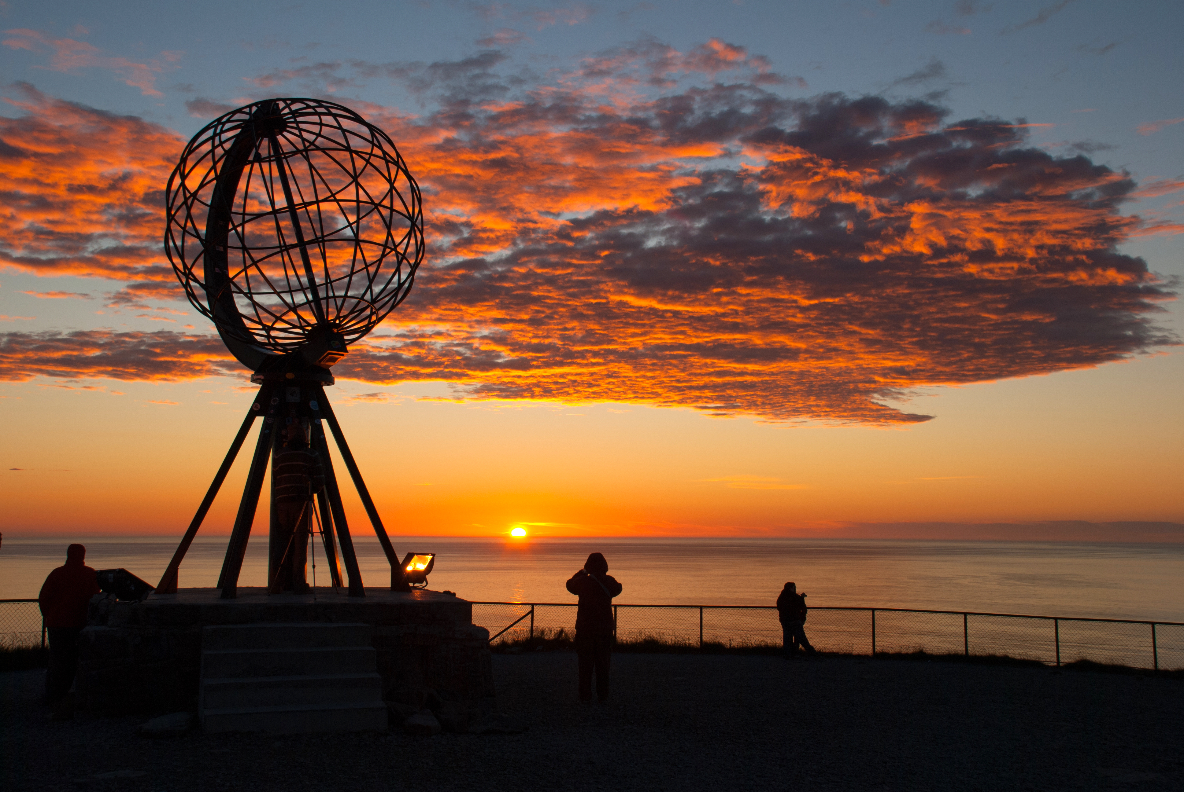 Fascinerende udsigt ved Nordkapps berømte globus-monument i Norge med silhuetter af rejsende mod den dramatiske midnatssol i den arktiske sommer