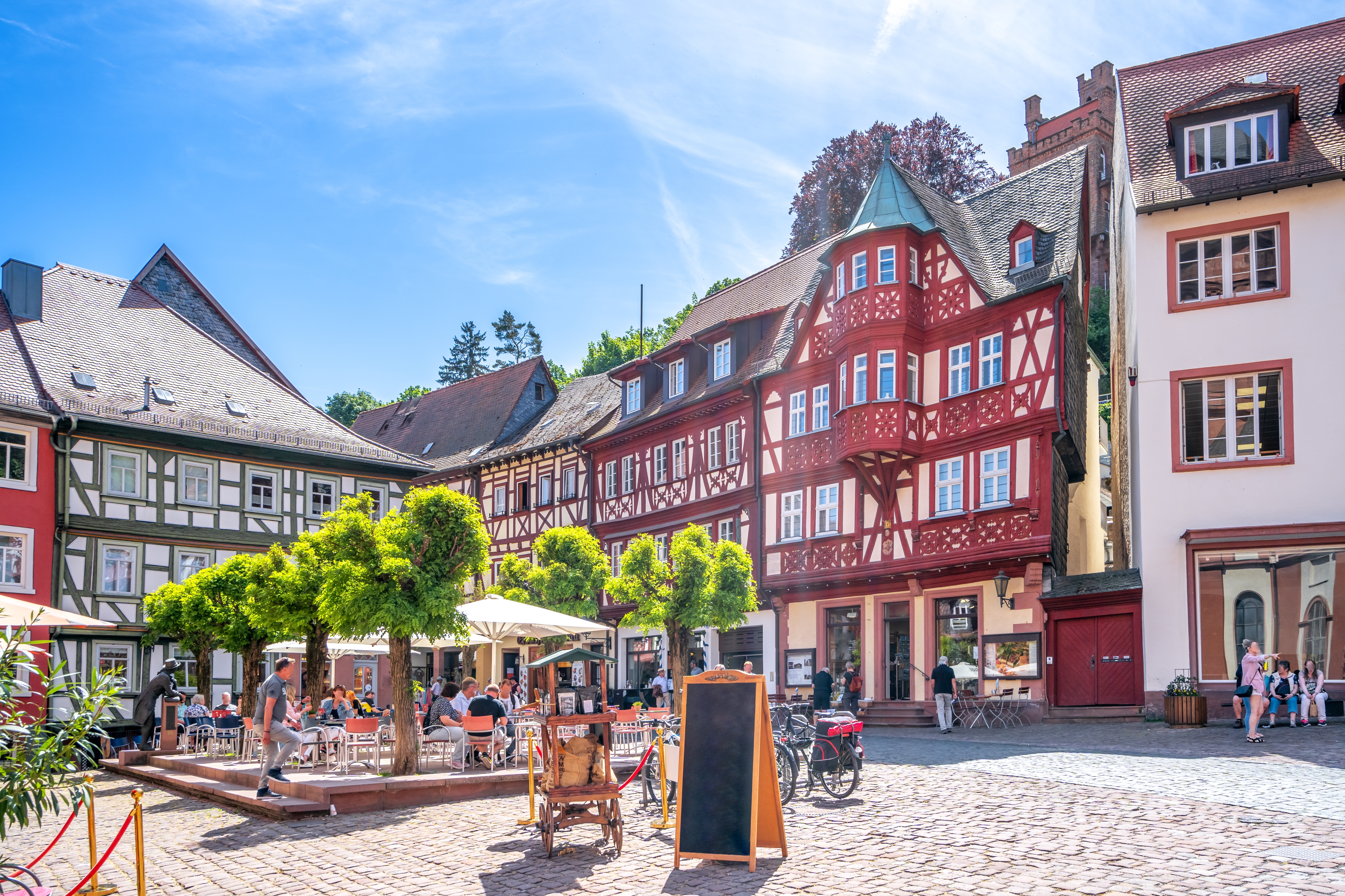Middelalderligt torv i Miltenberg, Tyskland, med maleriske bindingsværksbygninger, udeservering under store træer og stemningsfulde brolagte gader i sommervarmen