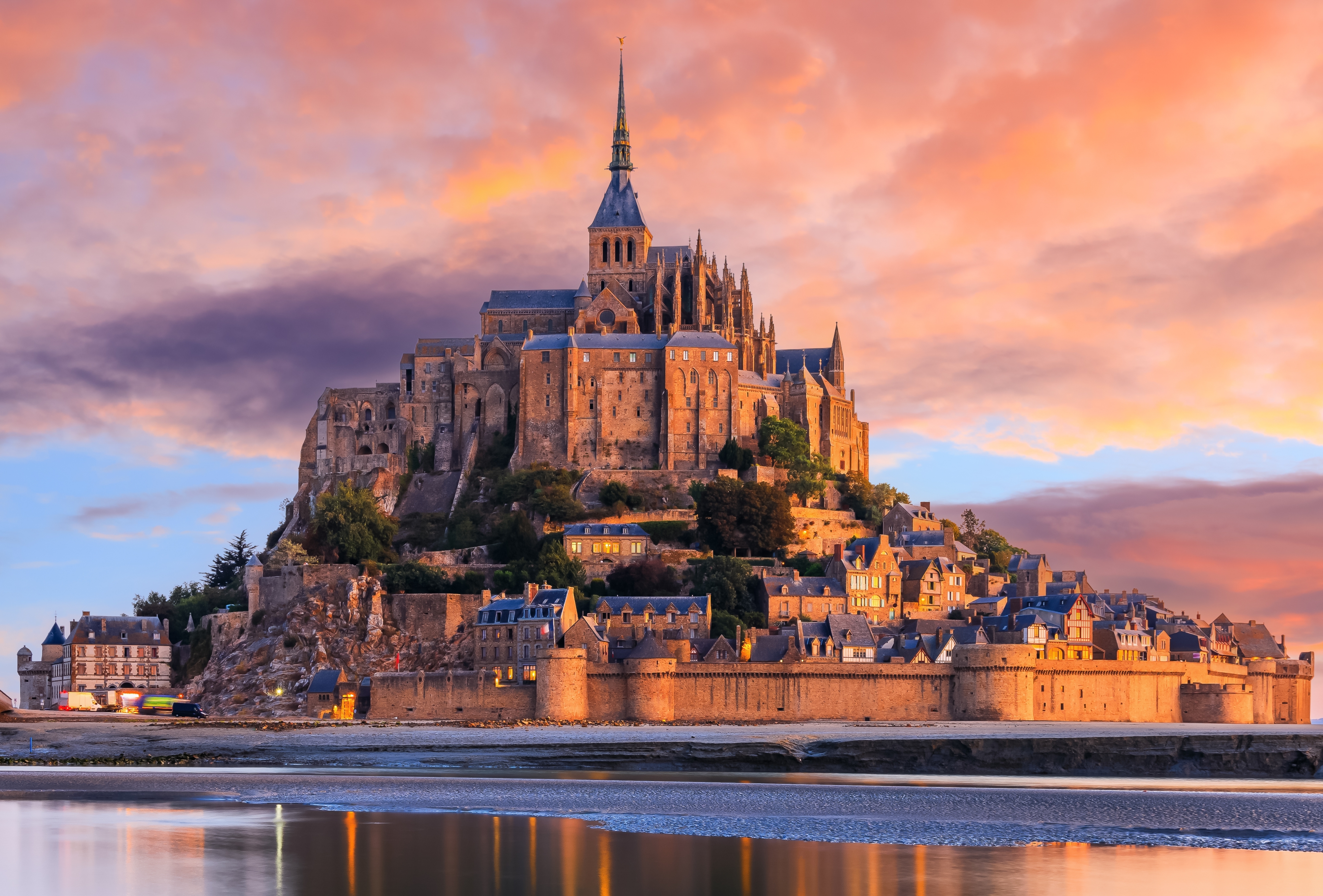 Mont Saint-Michel ved solopgang i Normandiet, Frankrig, med den smukke gotiske klosterkirke øverst på klippen og middelalderlige befæstninger langs vandkanten under en farverig himmel