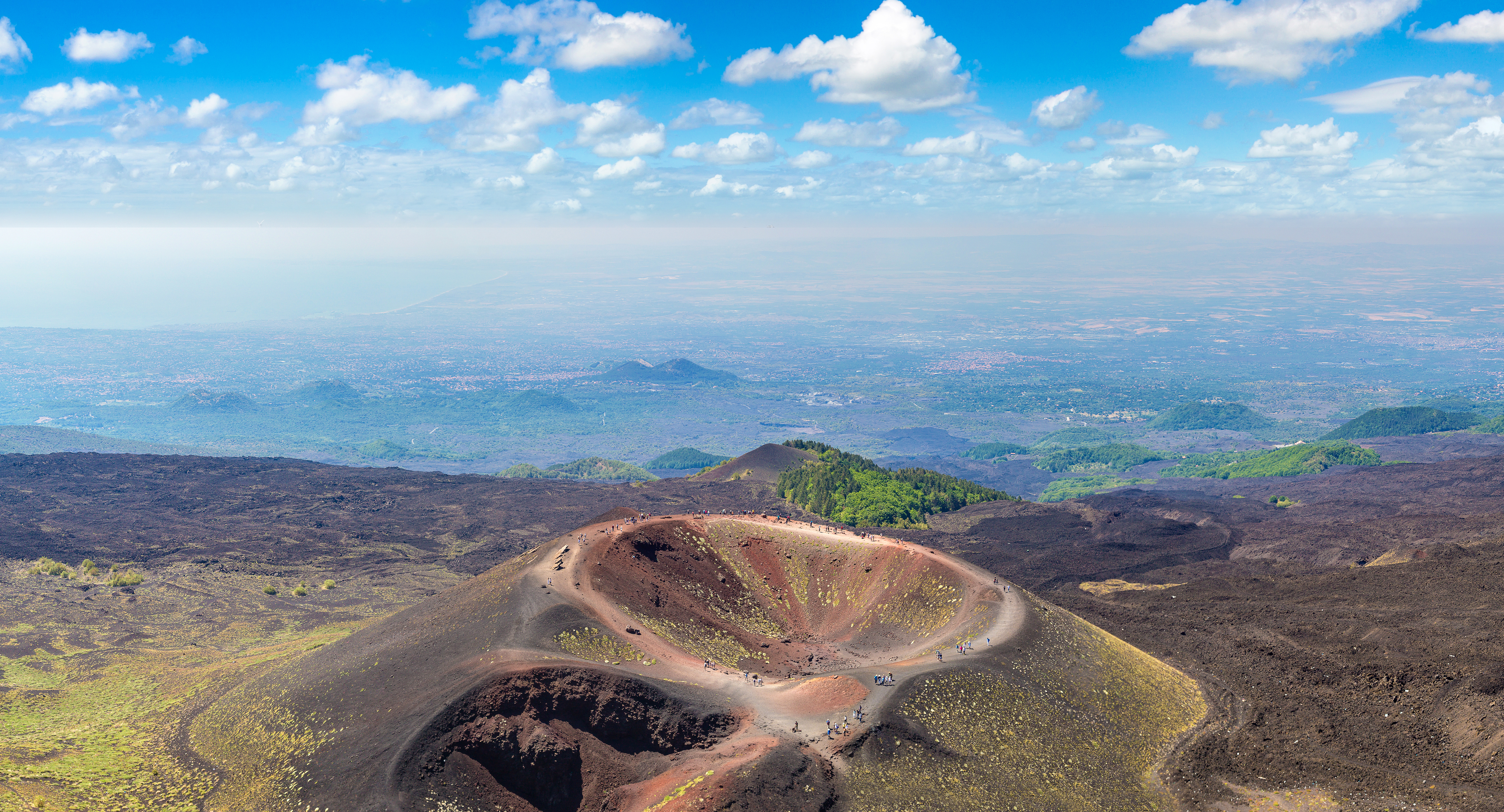 Luftfoto af den aktive vulkan Mount Etna på Sicilien med den imponerende kraterkant og det mørke lavalandskab omgivet af grøn vegetation og blå himmel