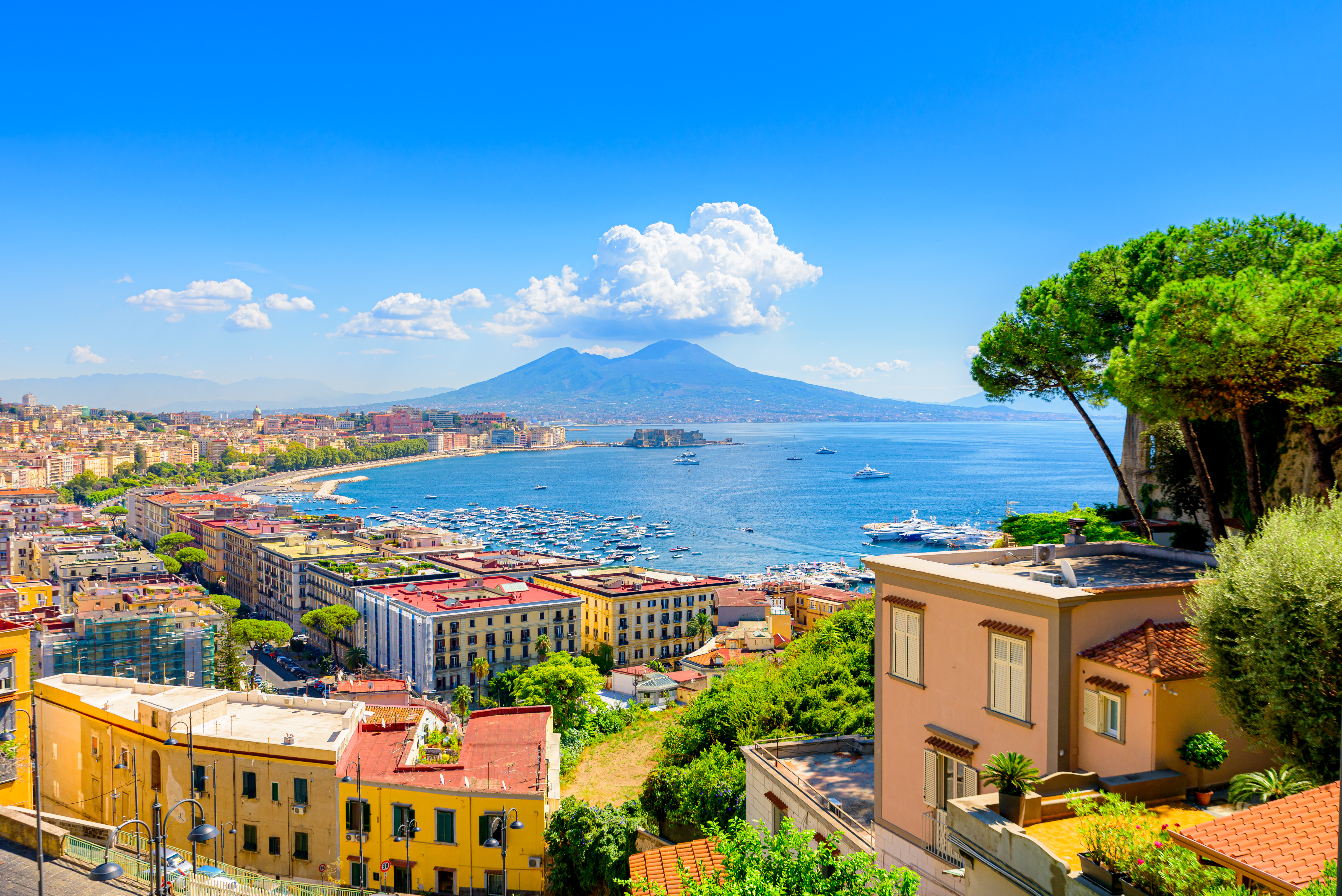 Pragtfuld panoramaudsigt over Napoli med den blå bugt, travl marina, historiske bygninger og Vesuv-vulkanen i baggrunden under en blå sommerhimmel