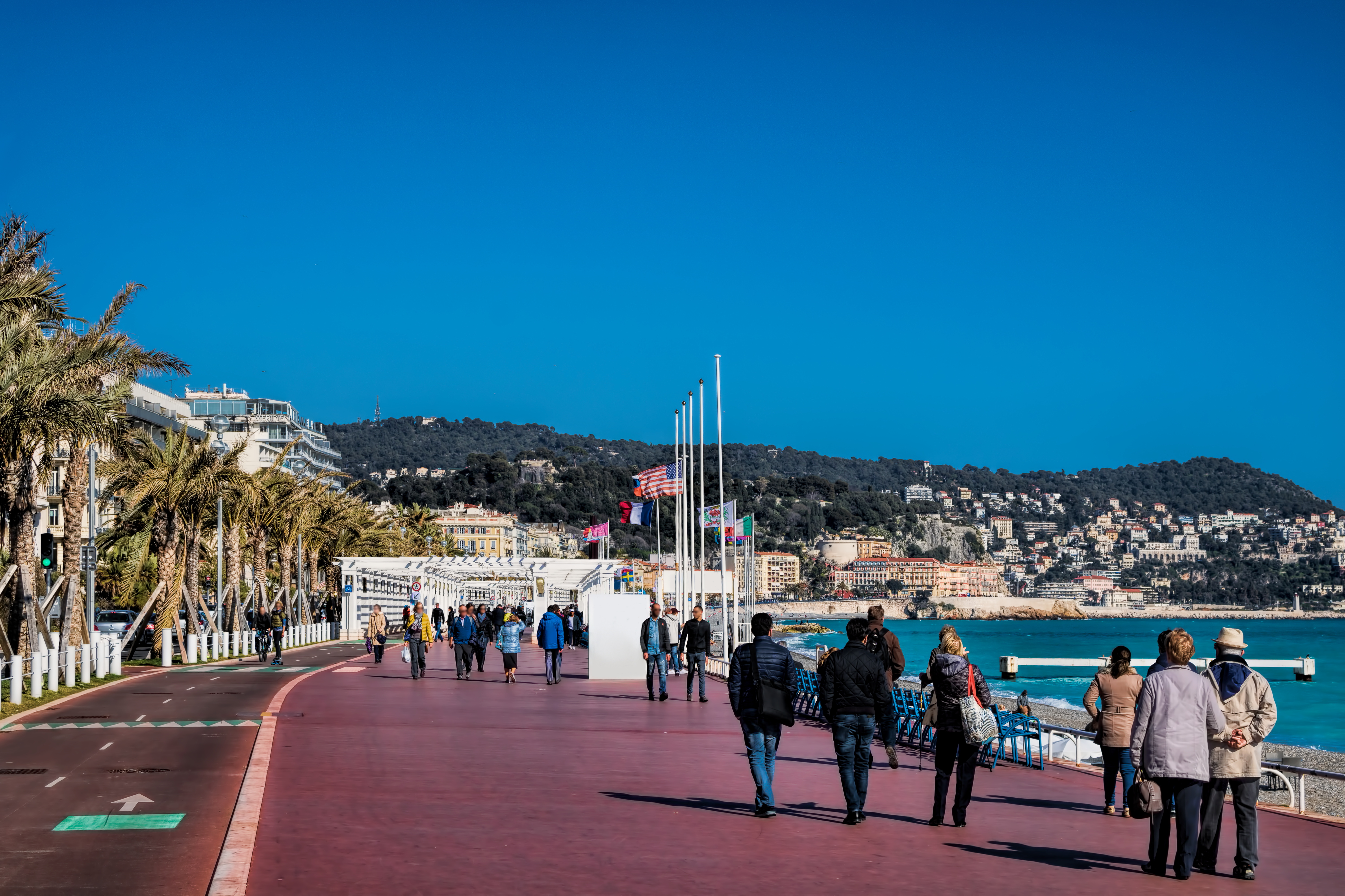 Gående turister på Promenade des Anglais i Nice med palmer og det smukke blå Middelhav i baggrunden