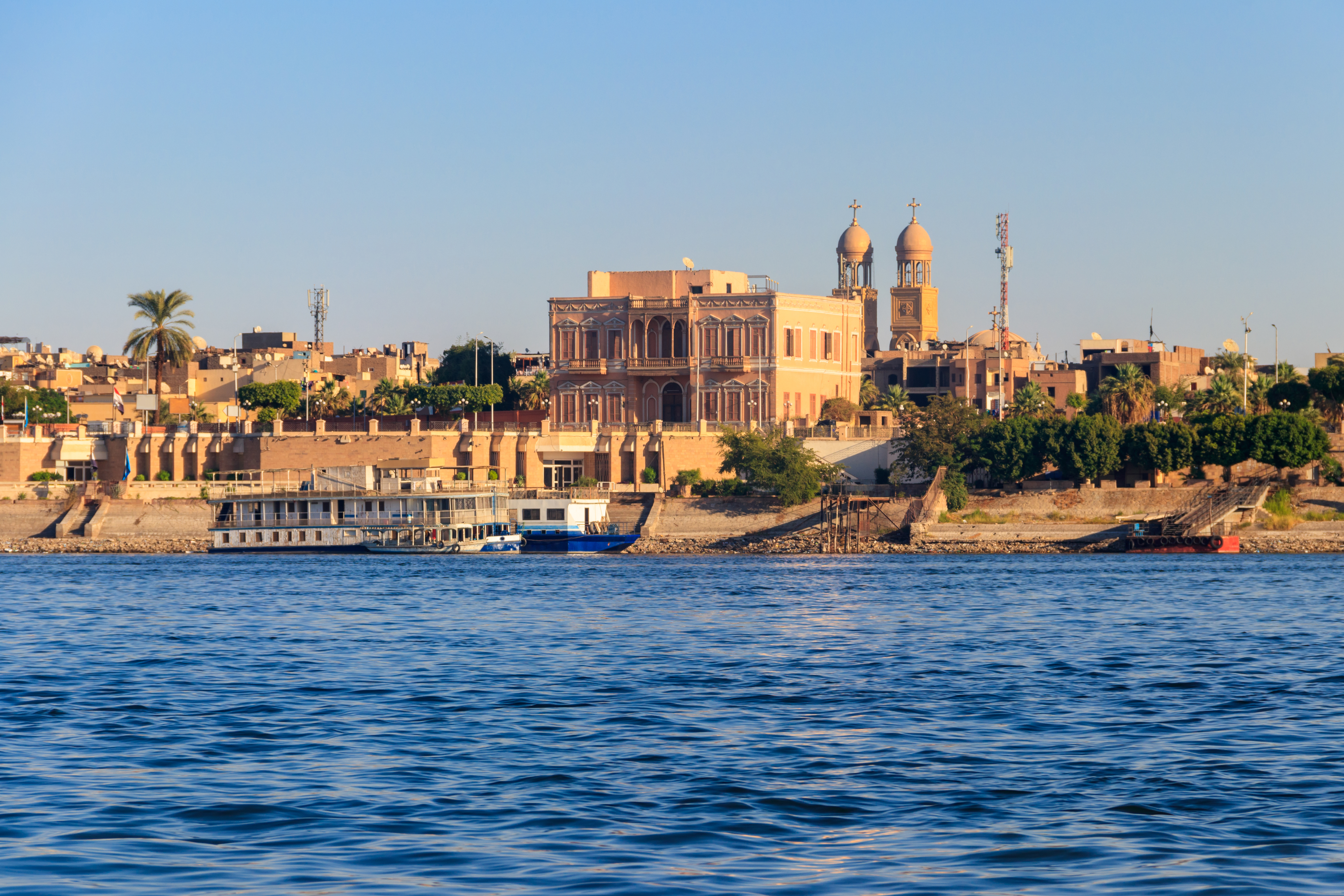Idyllisk udsigt over Luxor og Nilen med traditionelle bygninger, palmer og et krydstogtskib ved flodbredden under blå himmel