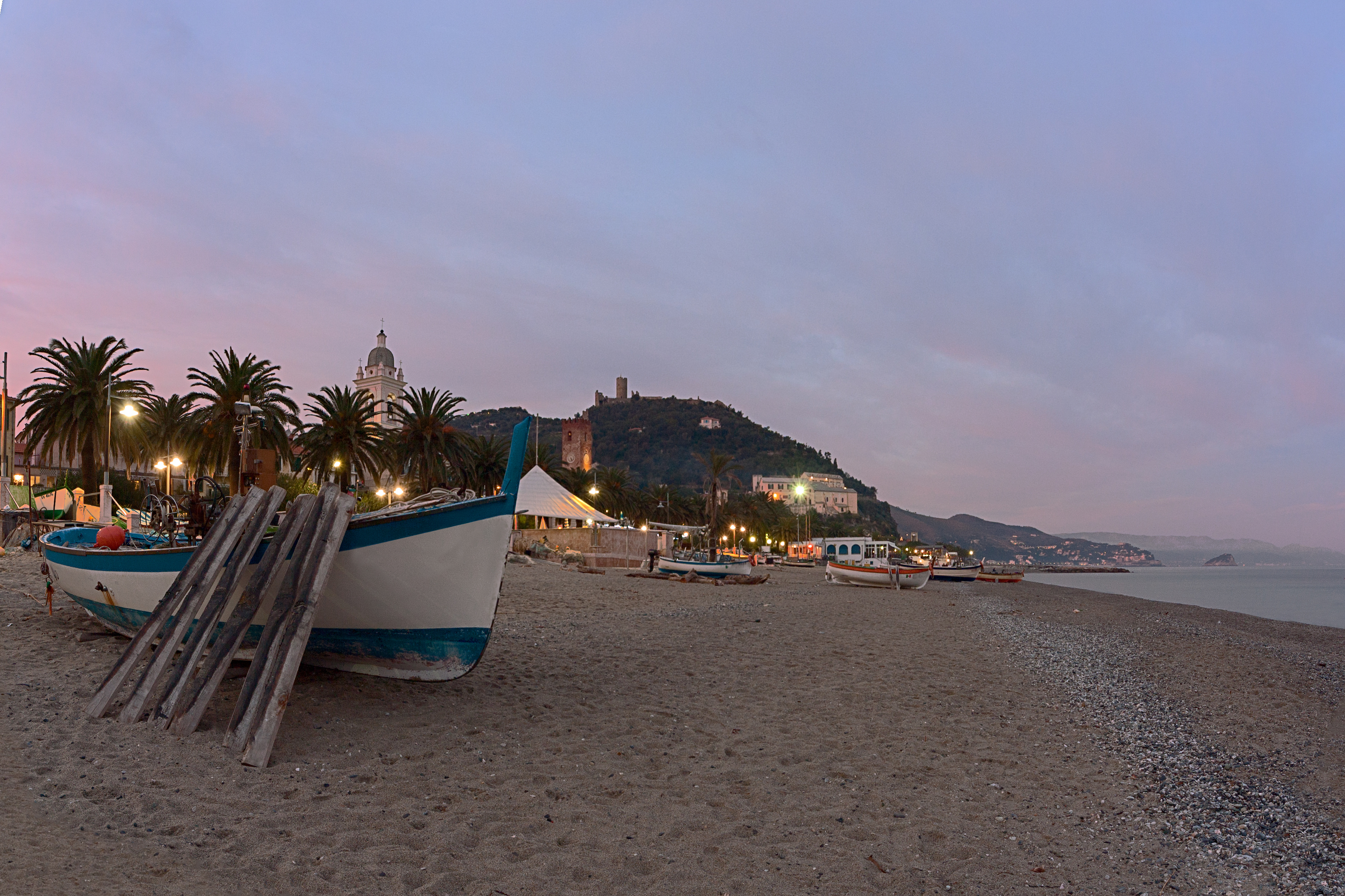 Hyggelig sandstrand i Noli med traditionel fiskerbåd ved solnedgang og palmetræer langs strandpromenaden i Ligurien, Italien