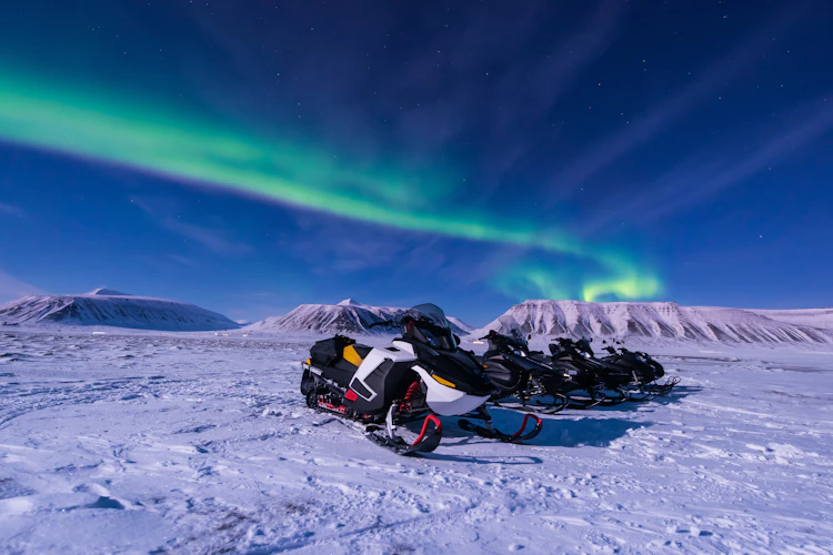 Nordlys danser hen over himlen mens snescootere holder parkeret i vinterlandskabet ved Longyearbyen på Svalbard