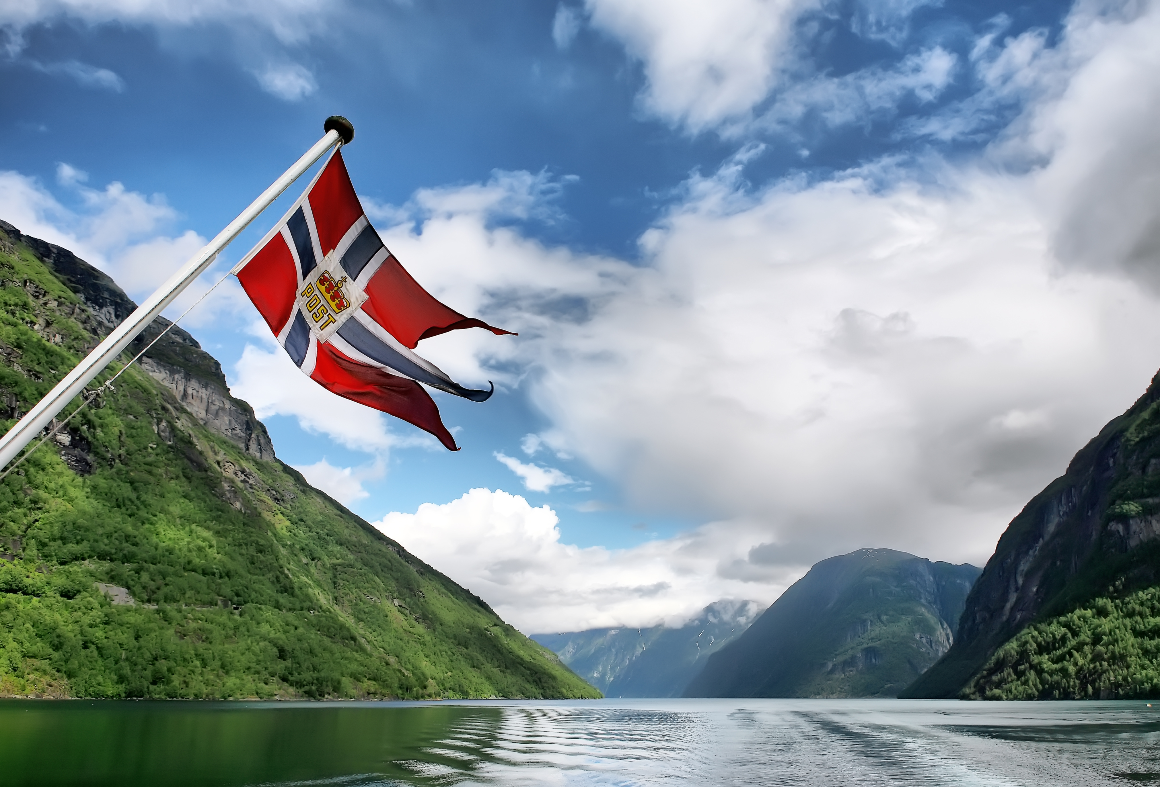 Norsk flag på en båd under sejltur gennem en idyllisk fjord med storslåede grønne bjerge og skyer på himlen i Norge