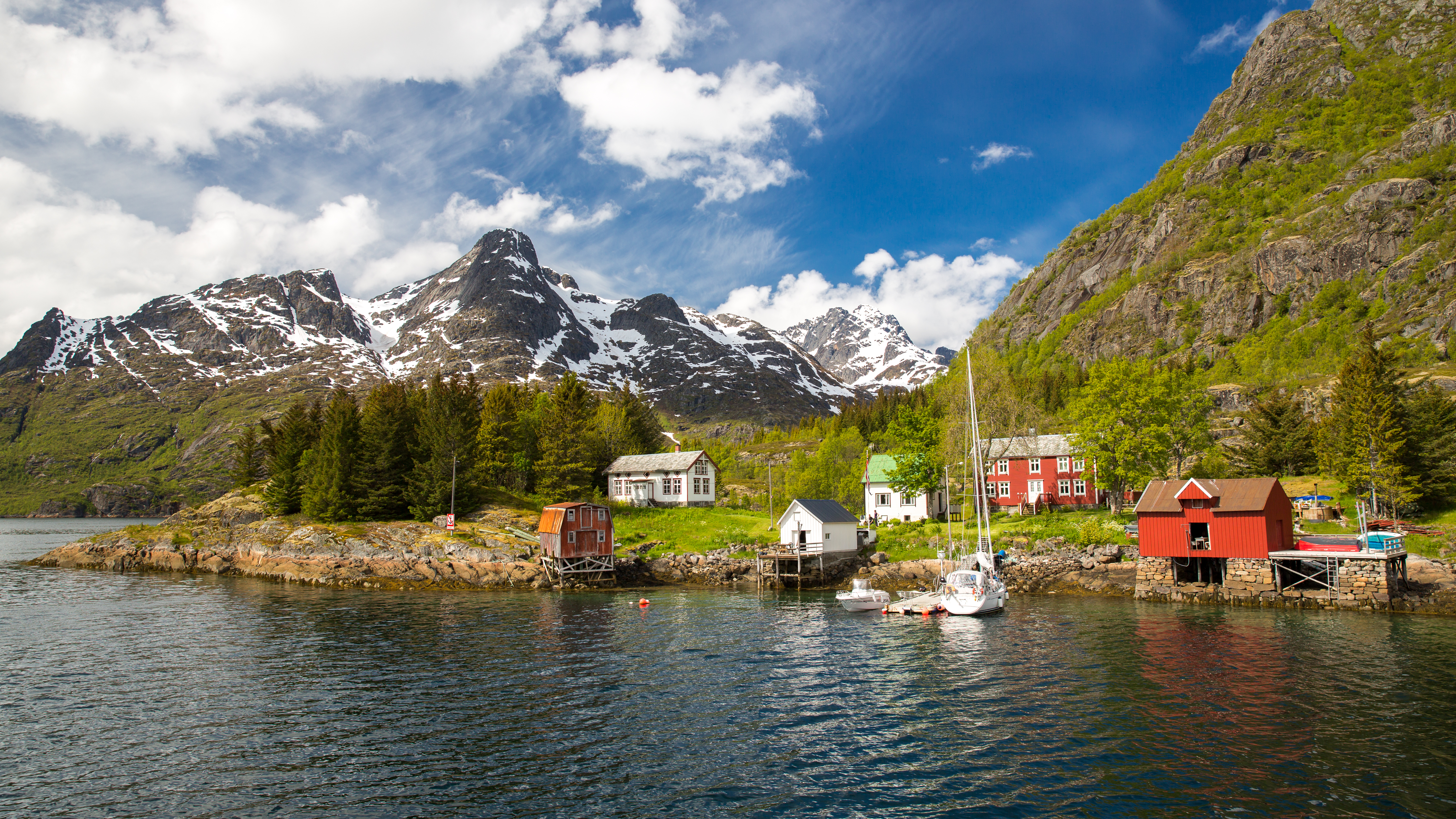 Naturskøn norsk fiskerihavn på Lofoten-øerne med røde huse, sejlbåde og dramatiske snedækkede fjelde under blå sommerhimmel