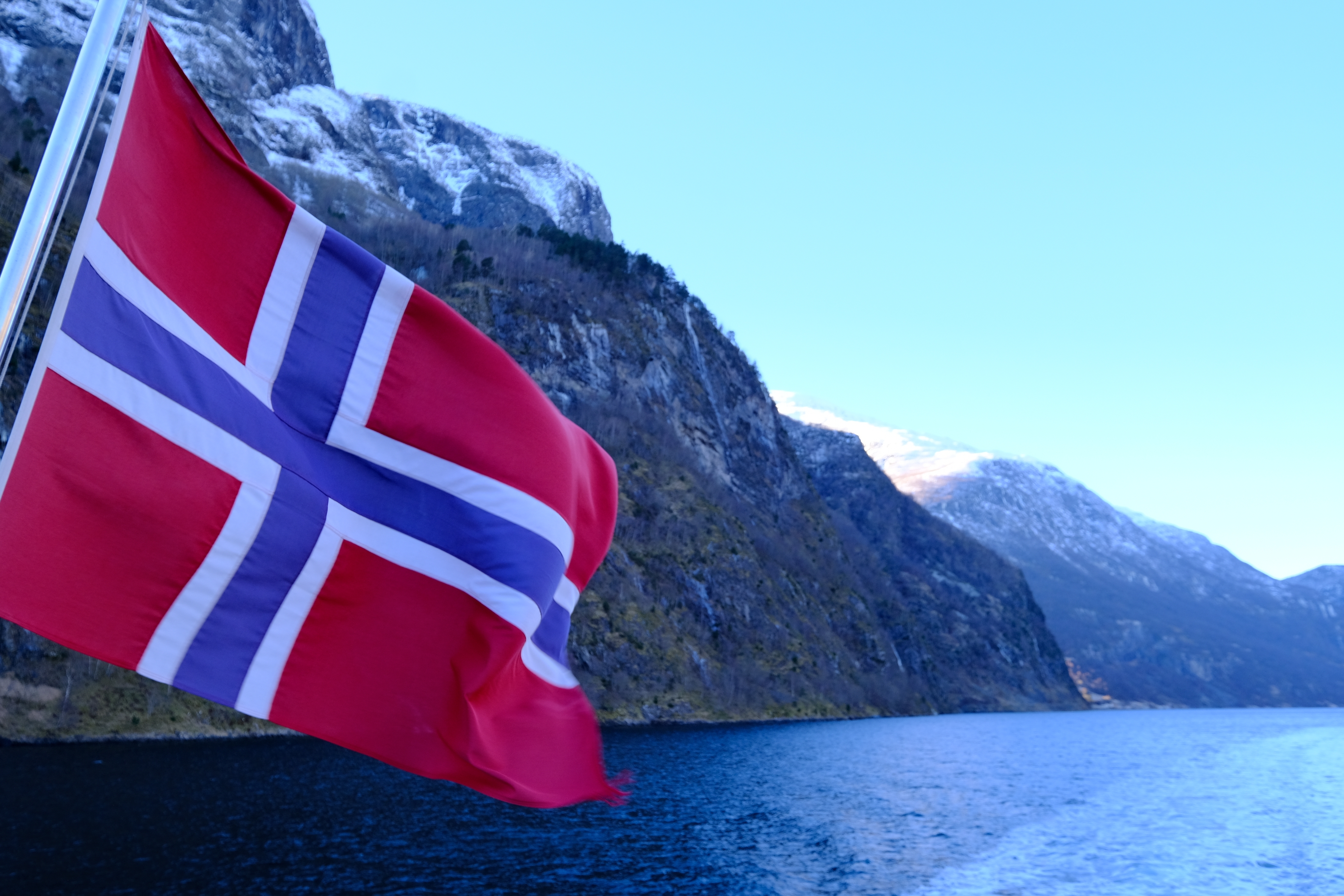 Norges flag vajer majestætisk over fjord omgivet af sneklædte bjergtinder