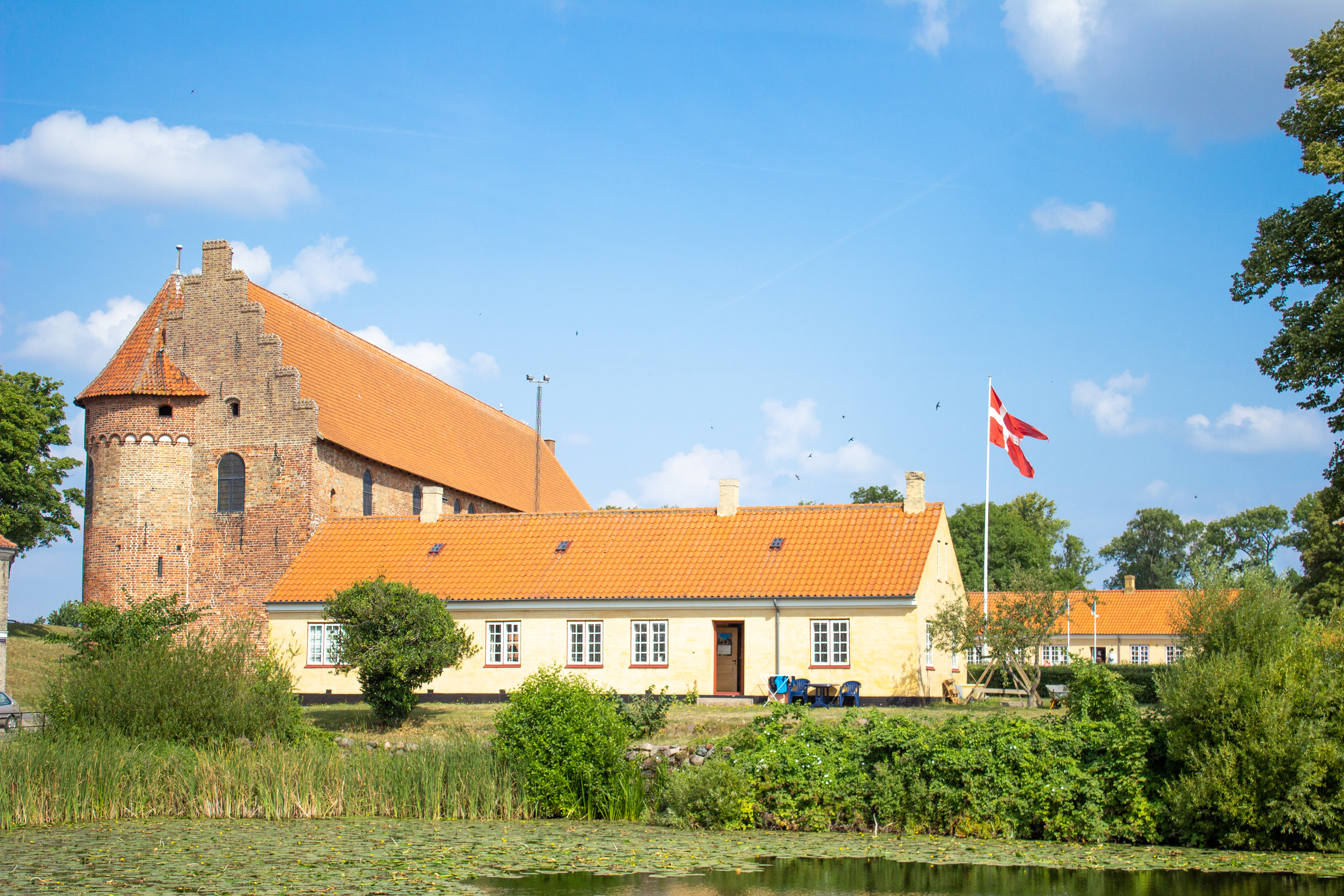 Idyllisk middelalderkirke med karakteristisk rundt tårn og orange tegltag spejlet i rolig sø i Nyborg