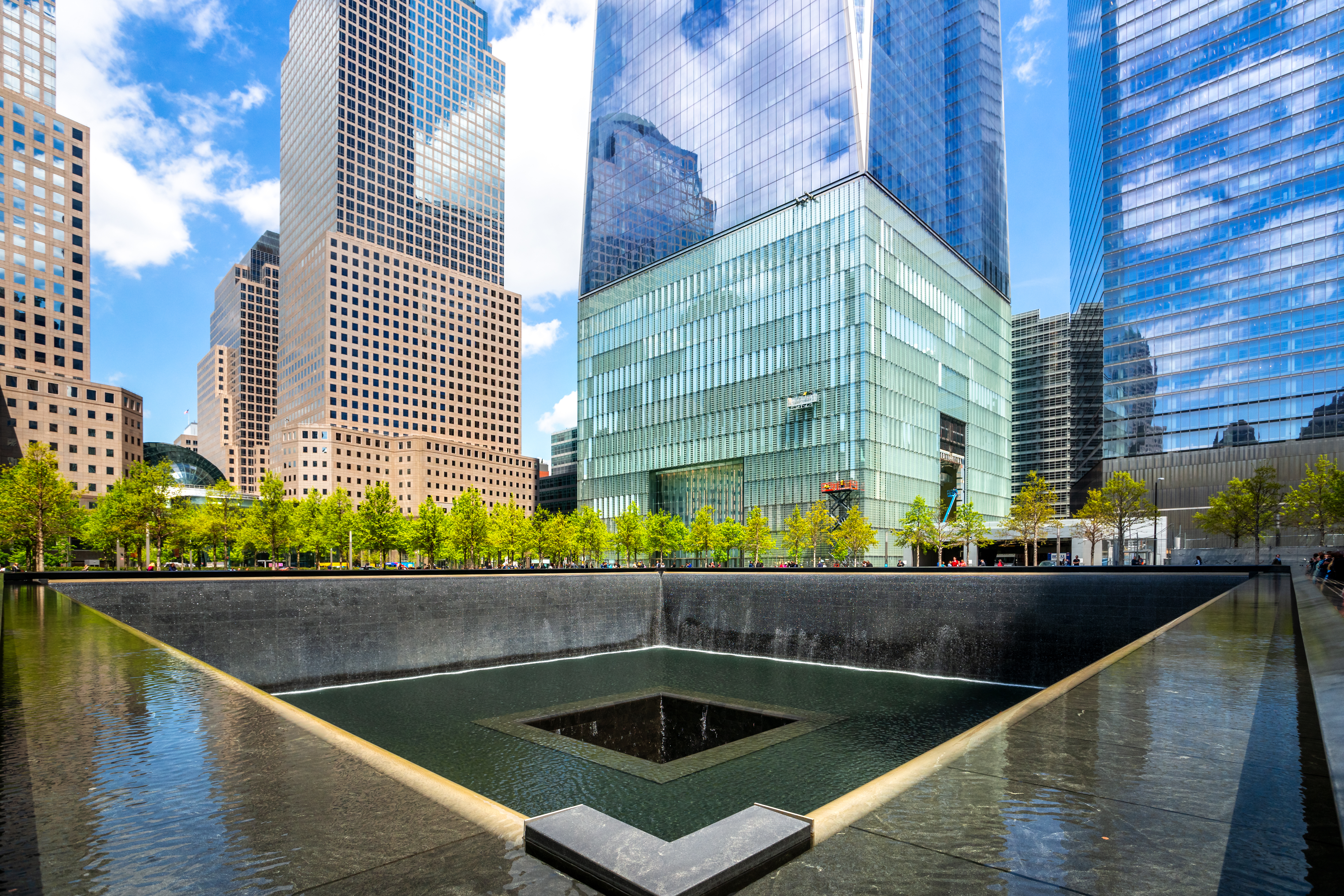 9/11 Memorialet i New York City med det store reflektionsbassin ved Ground Zero, omgivet af imponerende skyskrabere og grønne træer i sommersolen