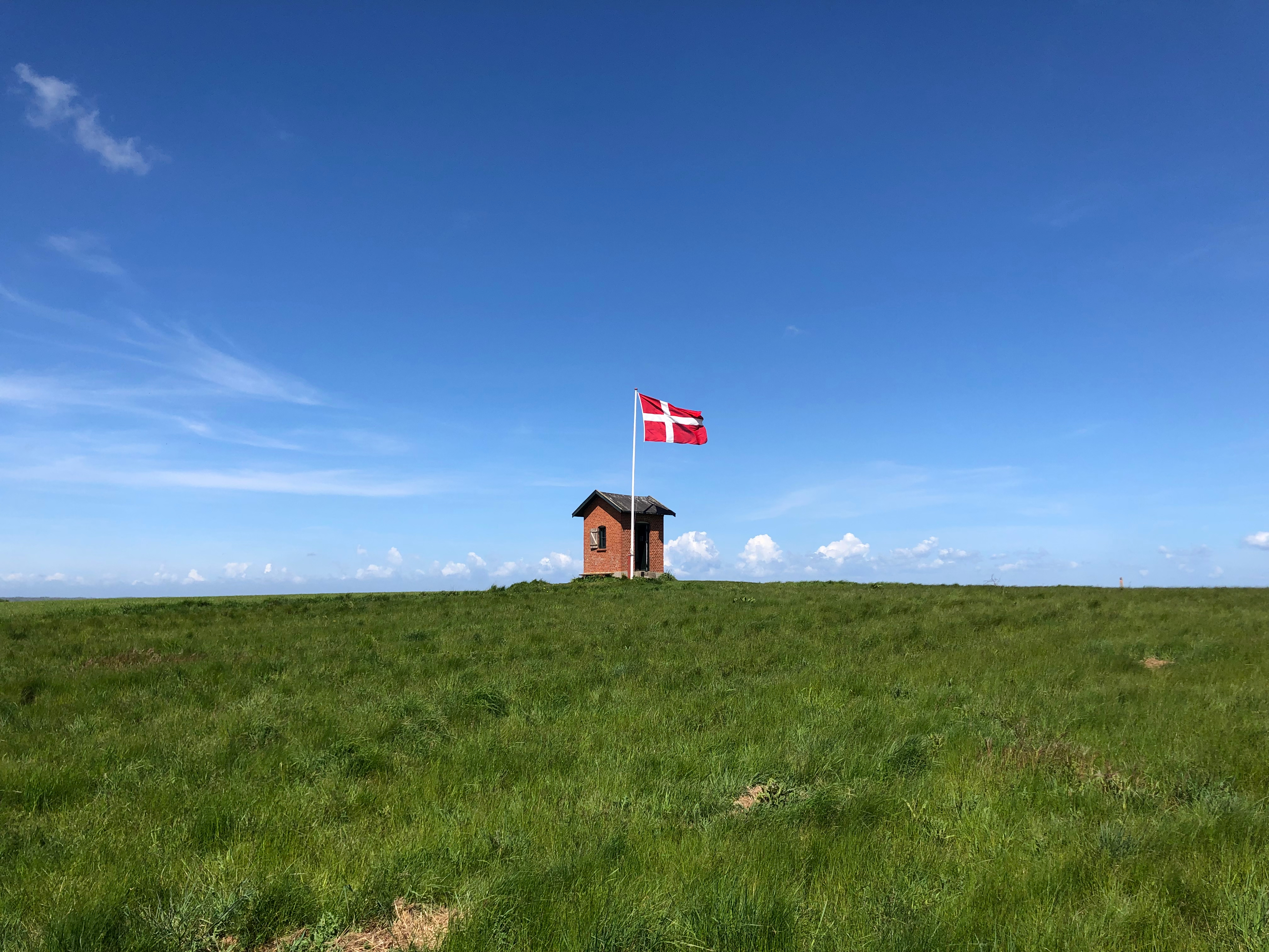 Lille rødt museum på Nyord med vajende Dannebrog og vid udsigt over grønne enge og blå sommerhimmel