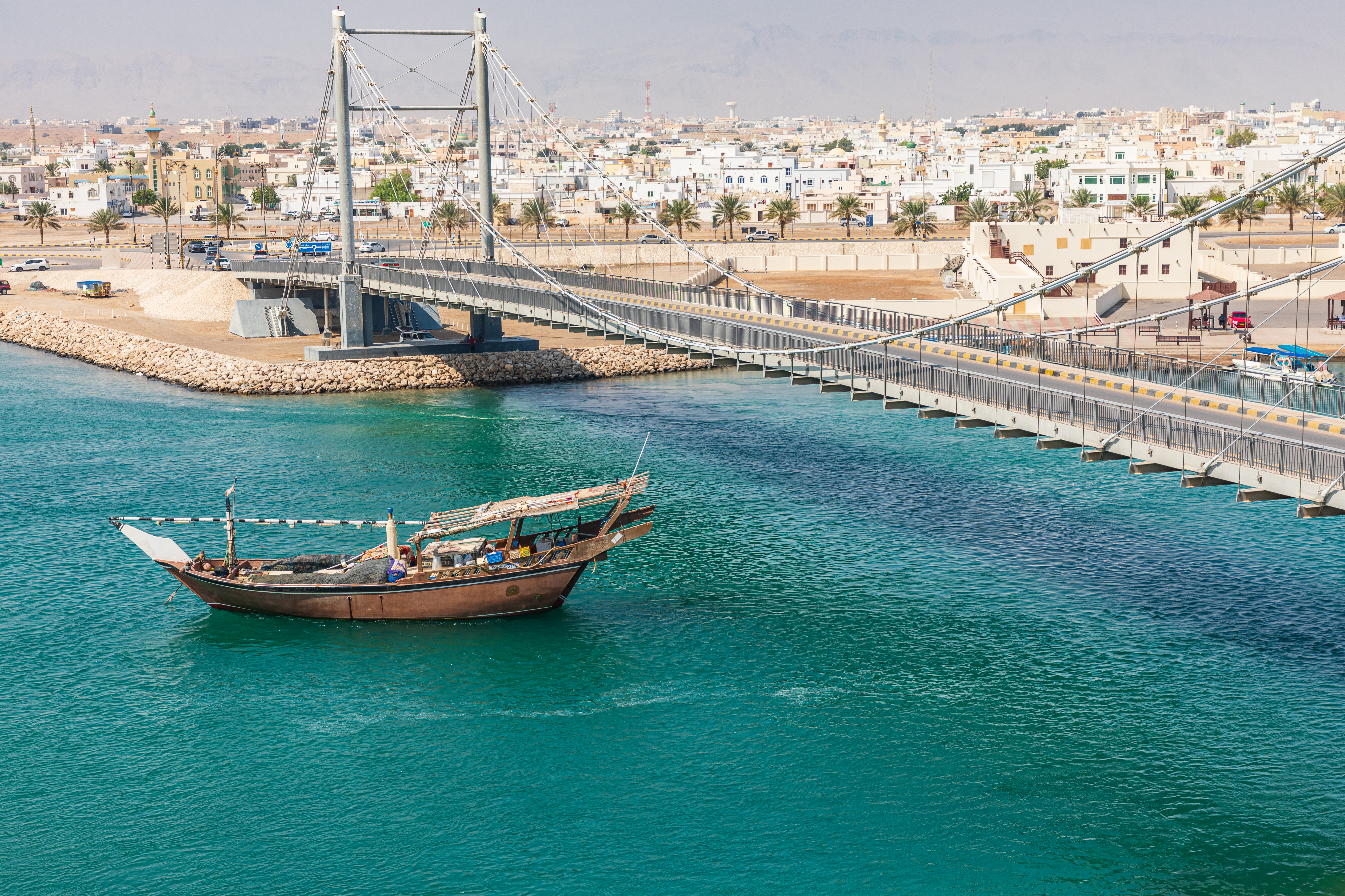 Traditionel omansk dhow i turkisblåt vand ved hængbroen i Sur, Oman, med byens hvide huse i baggrunden
