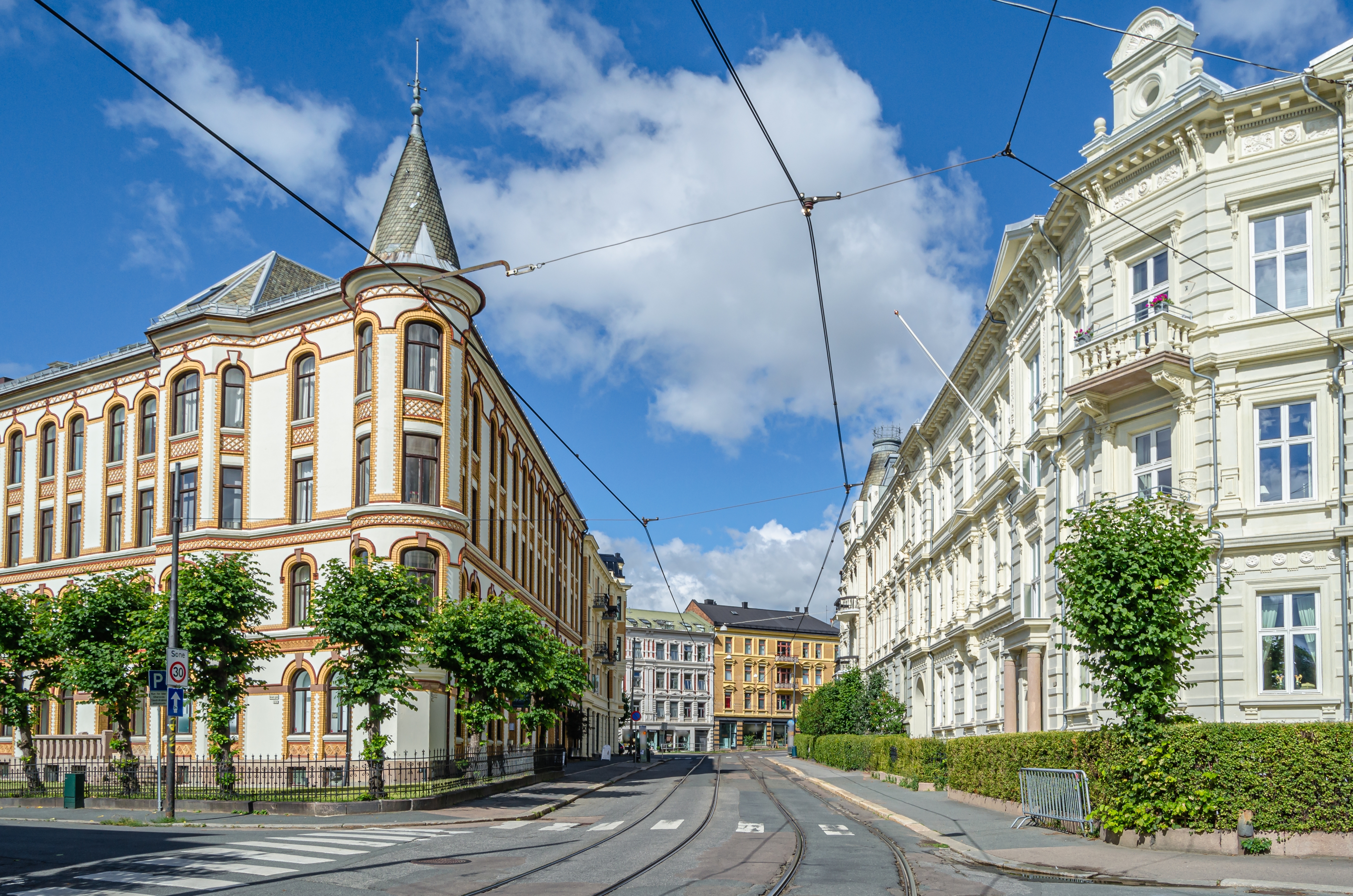 Imponerende jugendstil-bygninger langs en bred gade i Oslo, Norge, med frodig grøn beplantning og blå sommerhimmel