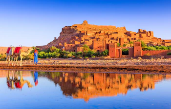 Smukt befæstet lerlandsby Ait Benhaddou ved Ouarzazate i Marokko med kameler og spejlbillede i floden ved solopgang