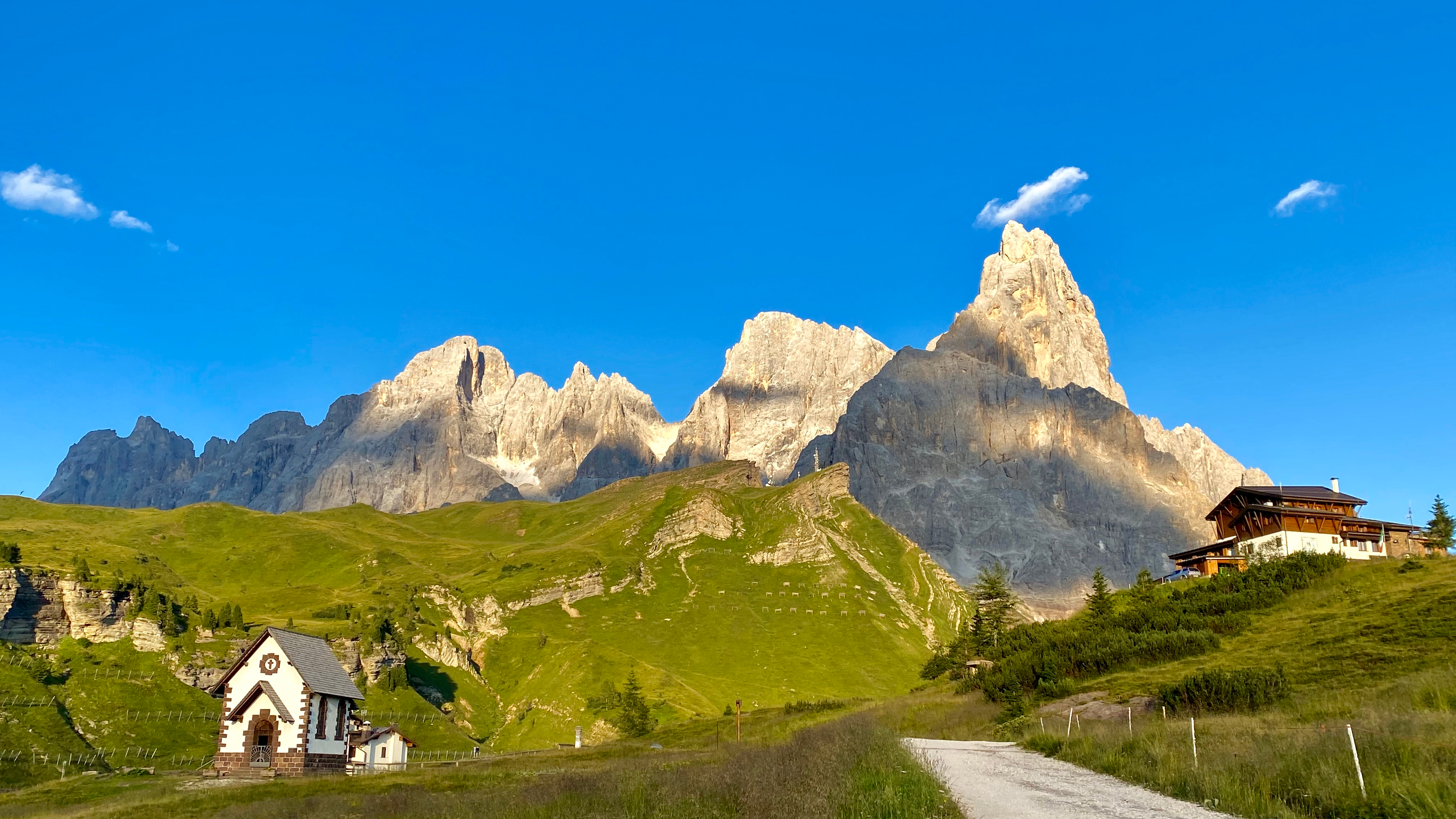 Fantastisk udsigt over Dolomitterne ved Passo Rolle med de ikoniske Pale di San Martino klippetinder, grønne alpenge og en charmerende bjergkirke i Trentino Alto Adige, Italien