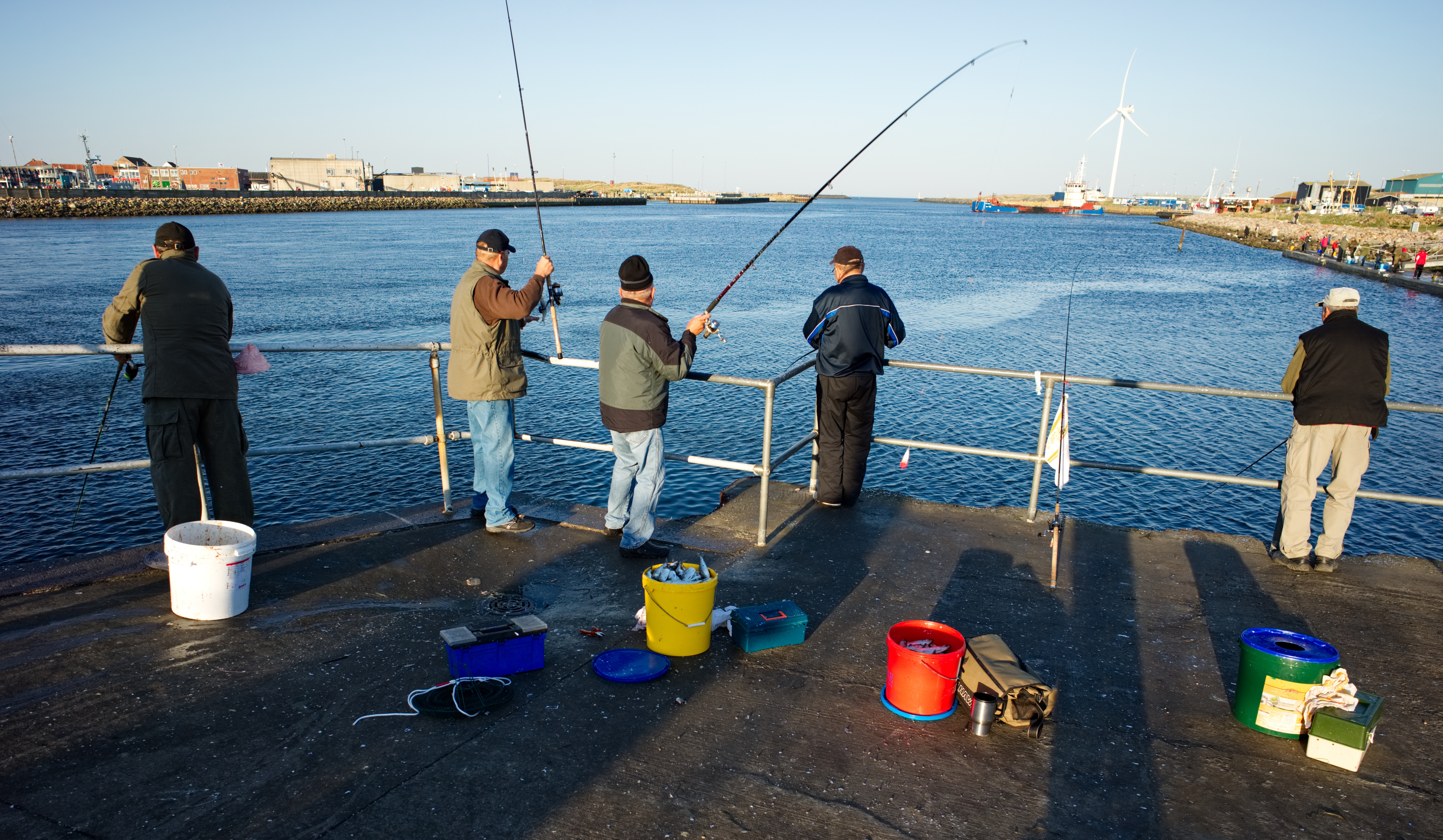 Mænd fisker fra kajen i Hvide Sande ved Nordsøen med bøjede fiskestænger og spande på betondækket