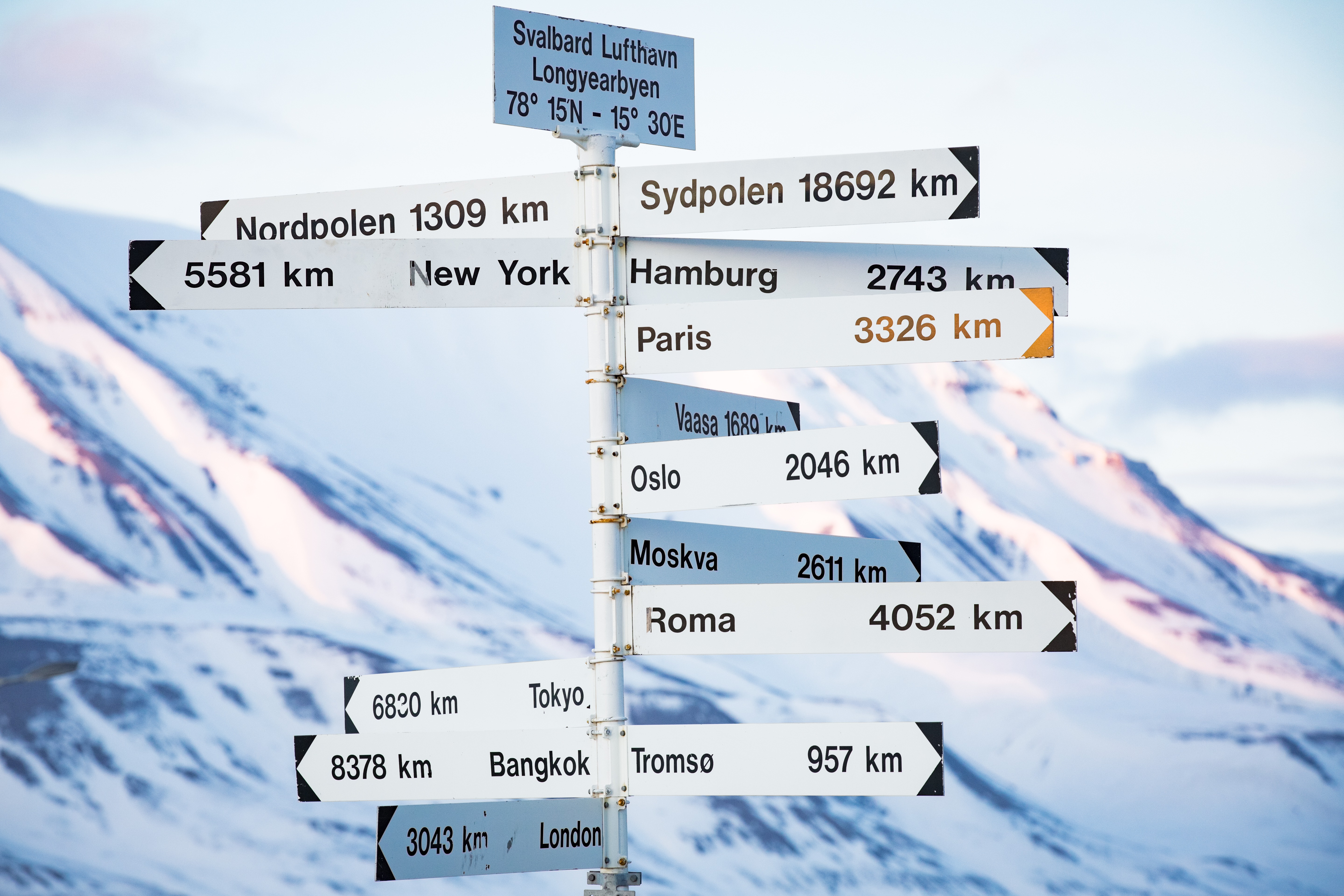 Polart destinationsguide skilt på Svalbard der angiver rejseafstande til verdenshovedstæder i et snedækket bjerglandskab