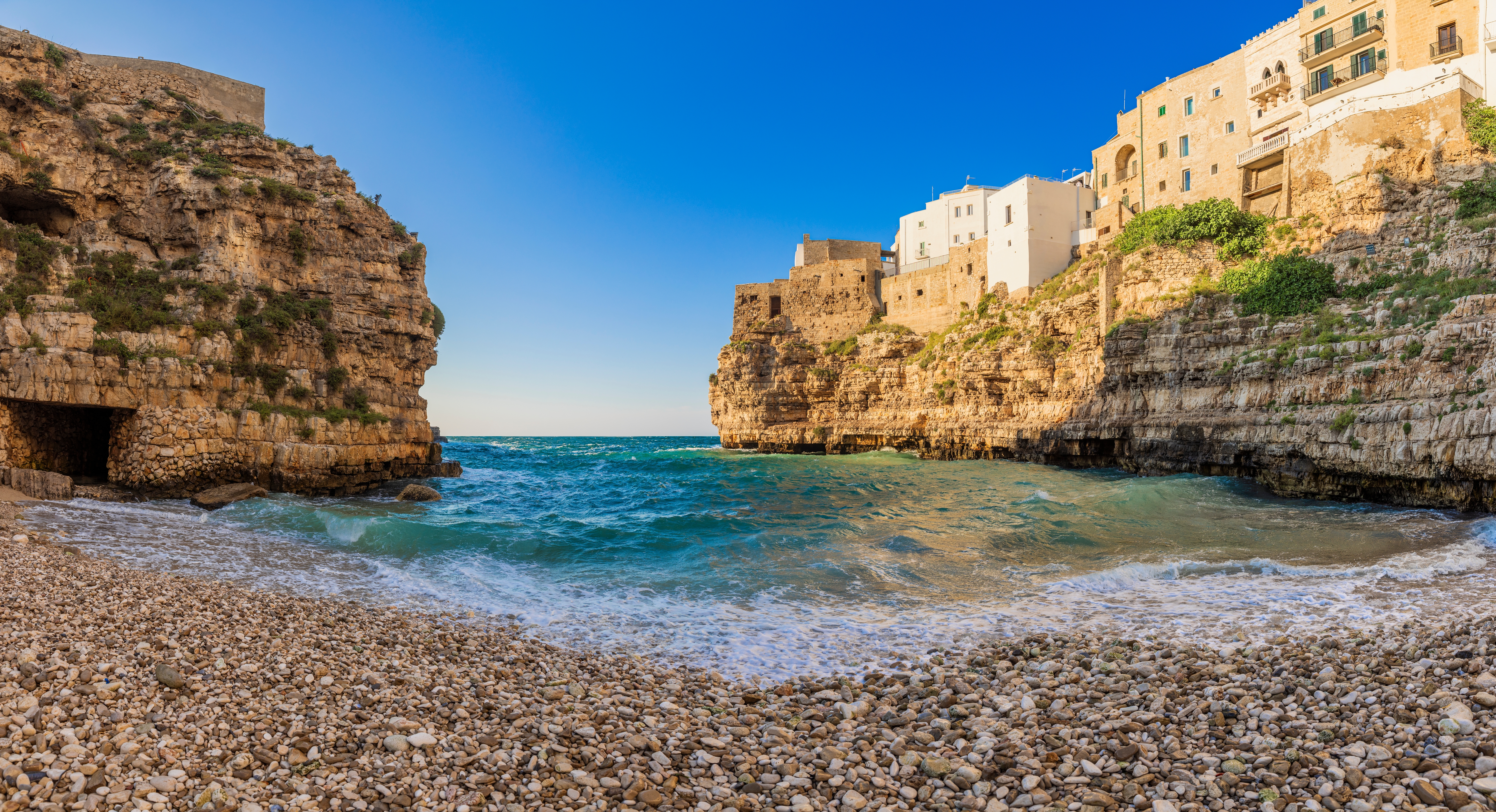 Fantastisk udsigt over Polignano a Mares stensstrand med det klare turkisblå Adriaterhav, omgivet af høje kalkstensklipper og charmerende hvide bygninger i Puglia, Italien