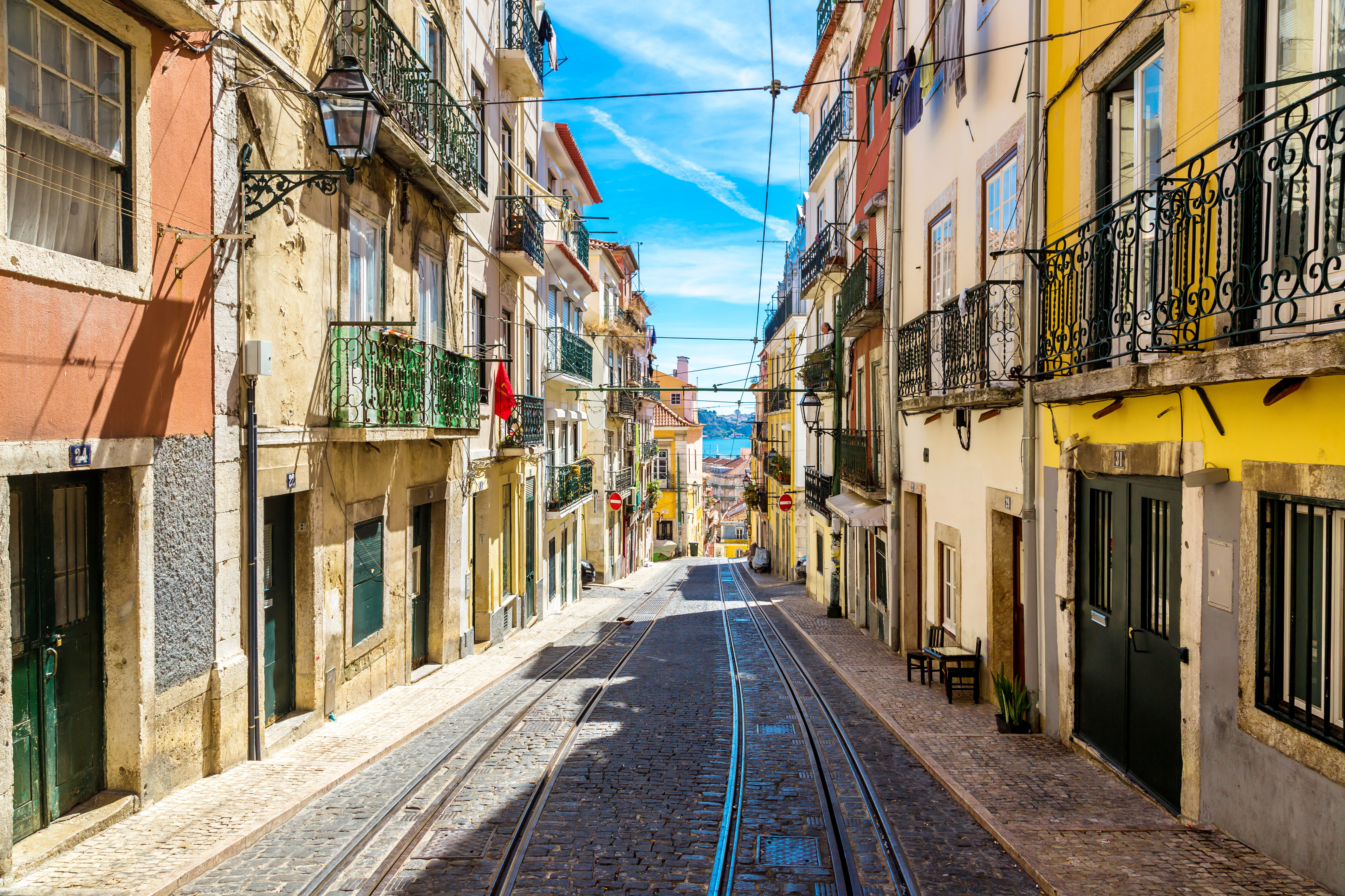 Stemningsfuld smal gade i det historiske Alfama-område i Lissabon med autentiske portugisiske huse, sporvognsskinner og den karakteristiske gule sporvogn under en klar blå sommerhimmel