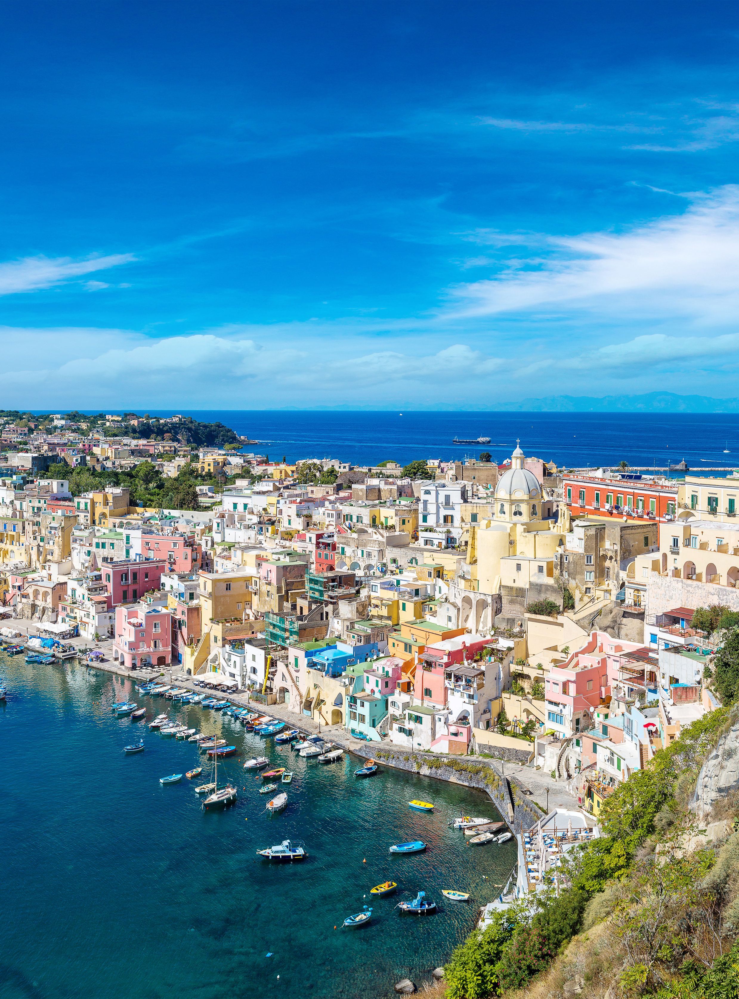Malerisk udsigt over den italienske ø Procida med pastellfarvede bygninger, azurblåt hav og fiskerbåde i den hyggelige havn under blå sommerhimmel