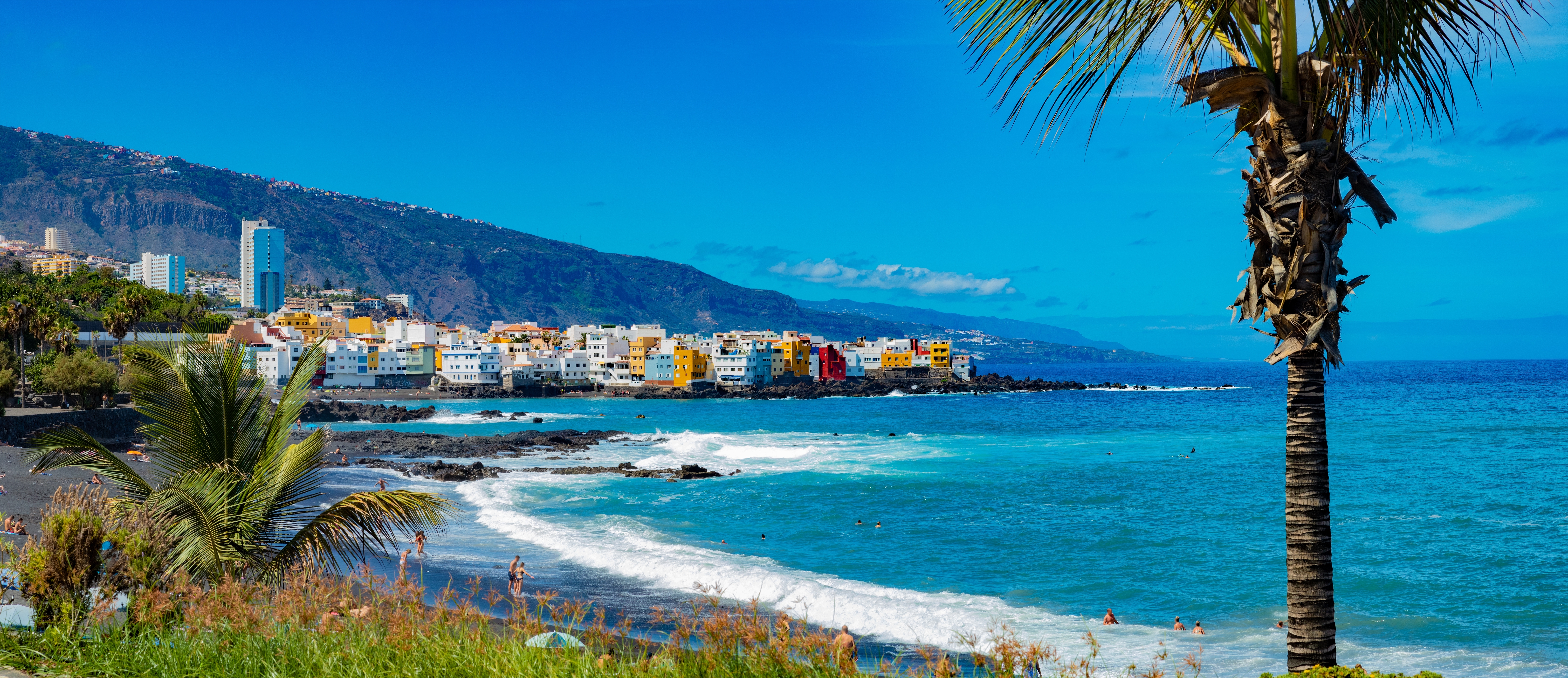 Solrig dag ved Puerto de la Cruz på Tenerife med bølger der slår mod kysten, palmer og farverige huse i baggrunden