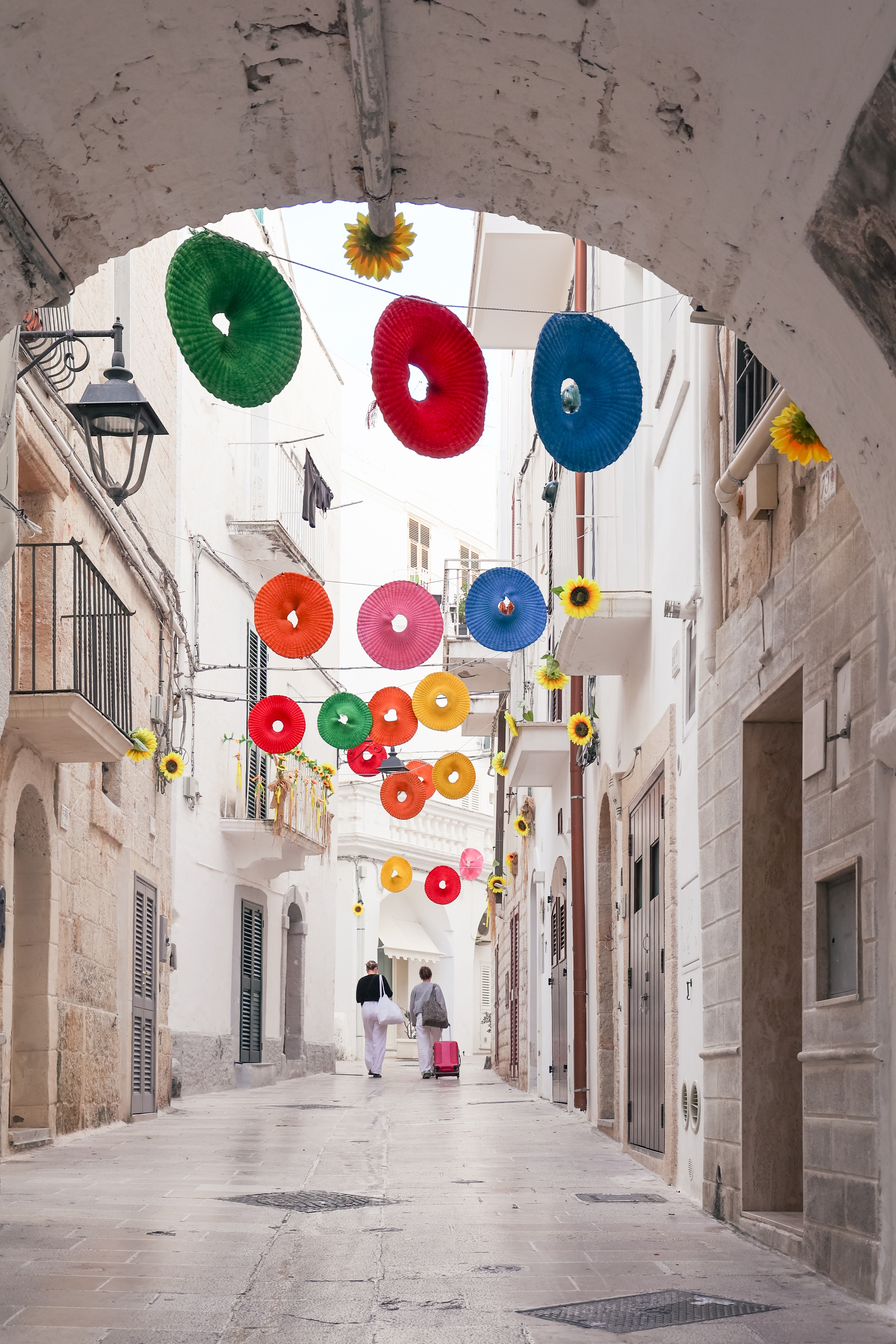 Turistpar går gennem en charmerende hvidkalket gade i Monopoli, Puglia med festlige farverige dekorationer hængende overhead