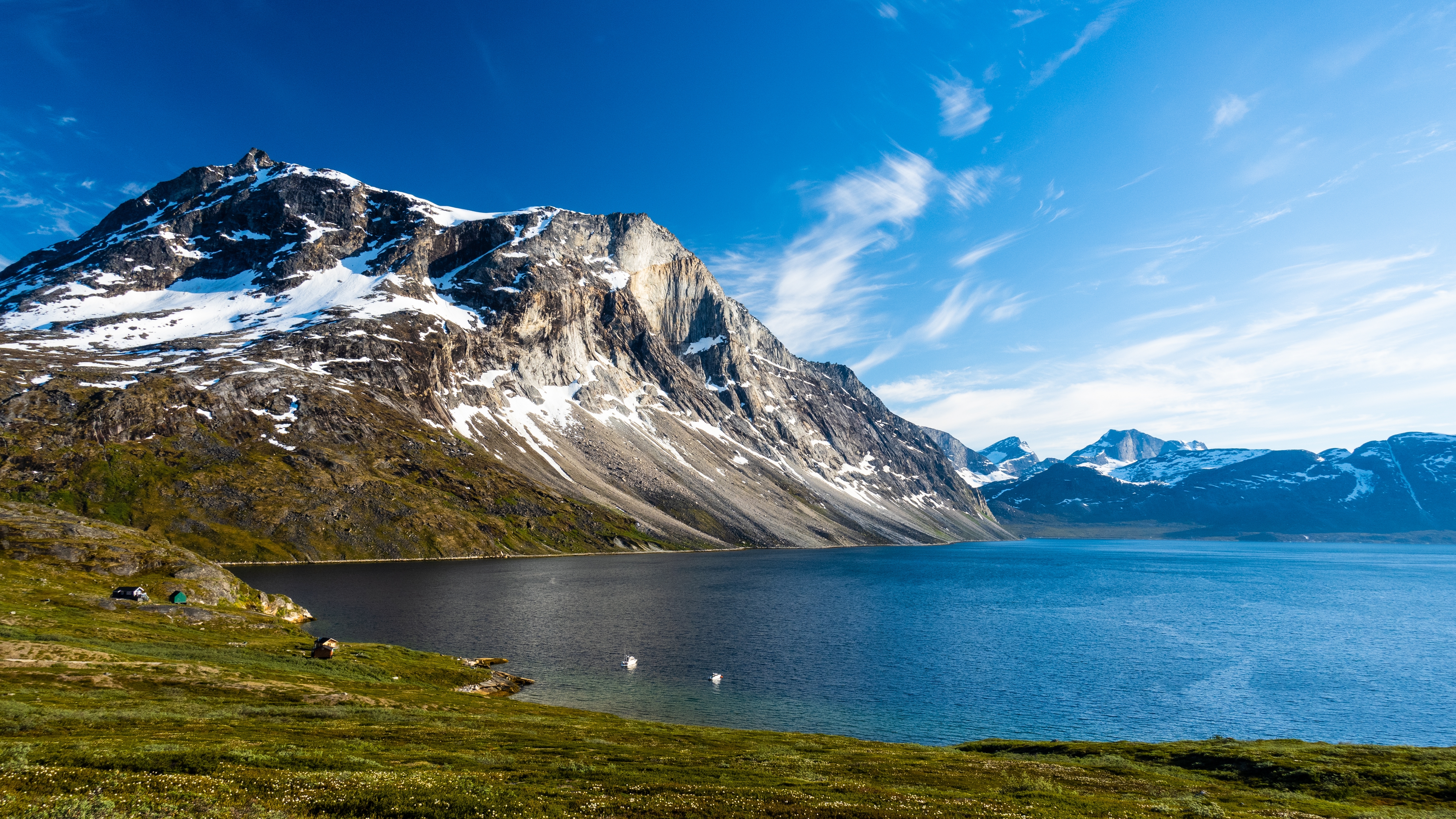 Eventyrligt nordisk landskab ved Qooqqut i Grønland med sneklædte bjerge, krystalklart blåt fjord og små hyggelige hytter langs den grønne kystlinje