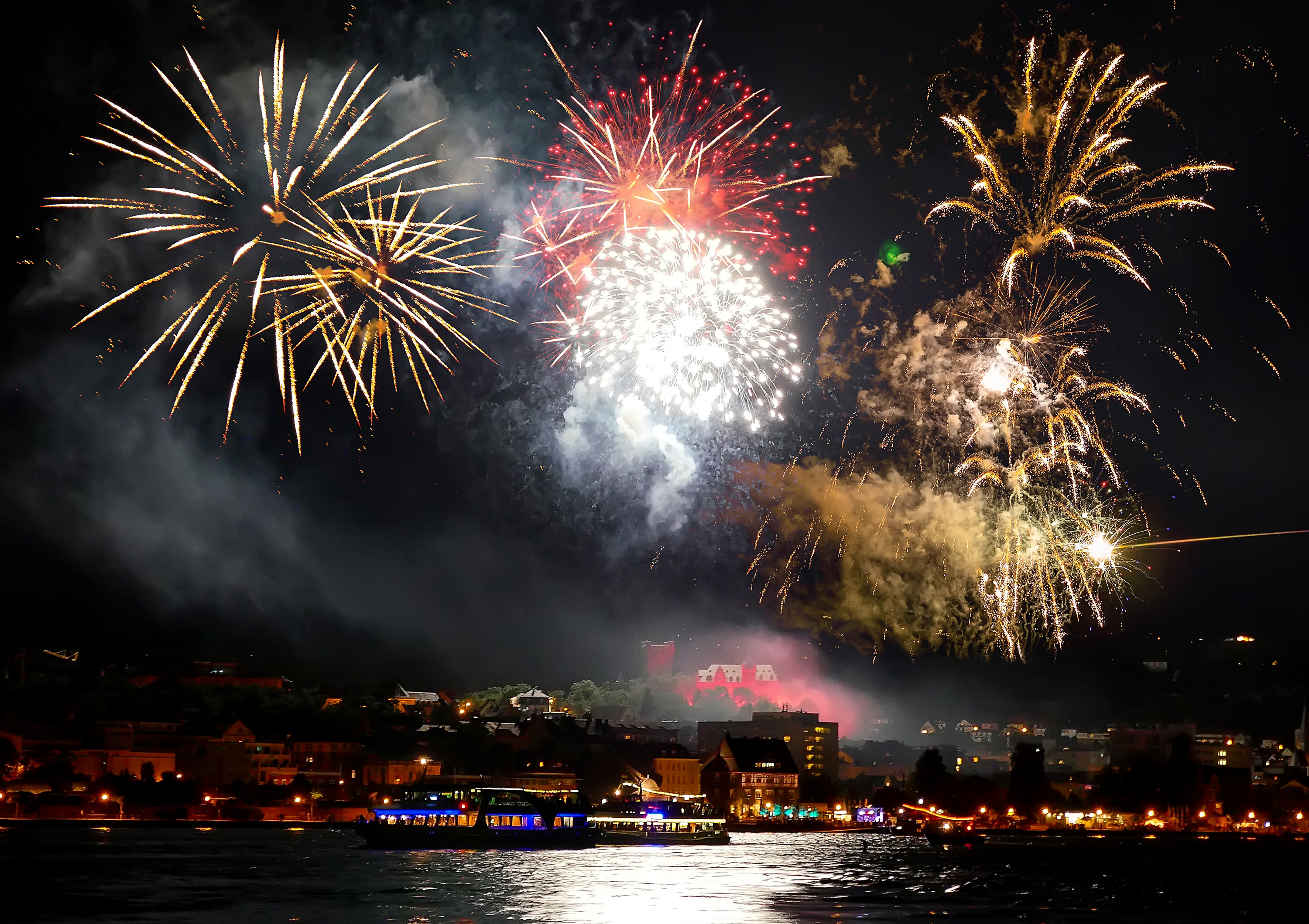 Imponerende fyrværkerioptog langs Rhinen ved Rüdesheim med farvestrålende eksplosioner i rødt og guld under Rhein in Flammen festivalen