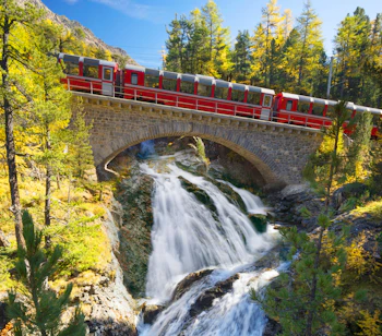Rødt alpetog på gammel stenbro over vandfald i schweiziske bjerge om efteråret