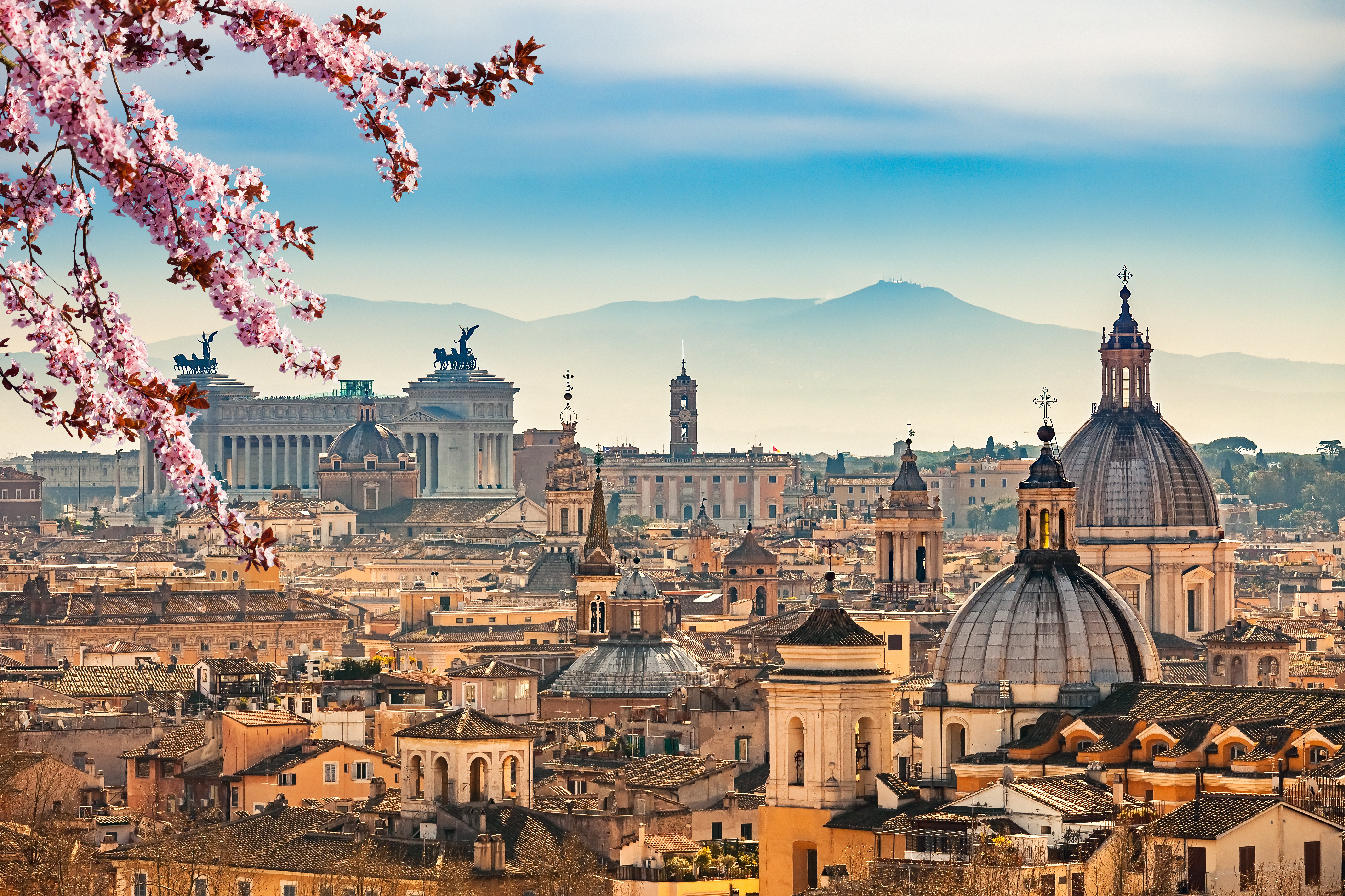 Storslået forårsudsigt over Rom med blomstrende kirsebærtræer, historiske monumenter og den ikoniske Vittoriano i gylden belysning, set fra det historiske Castel Sant'Angelo