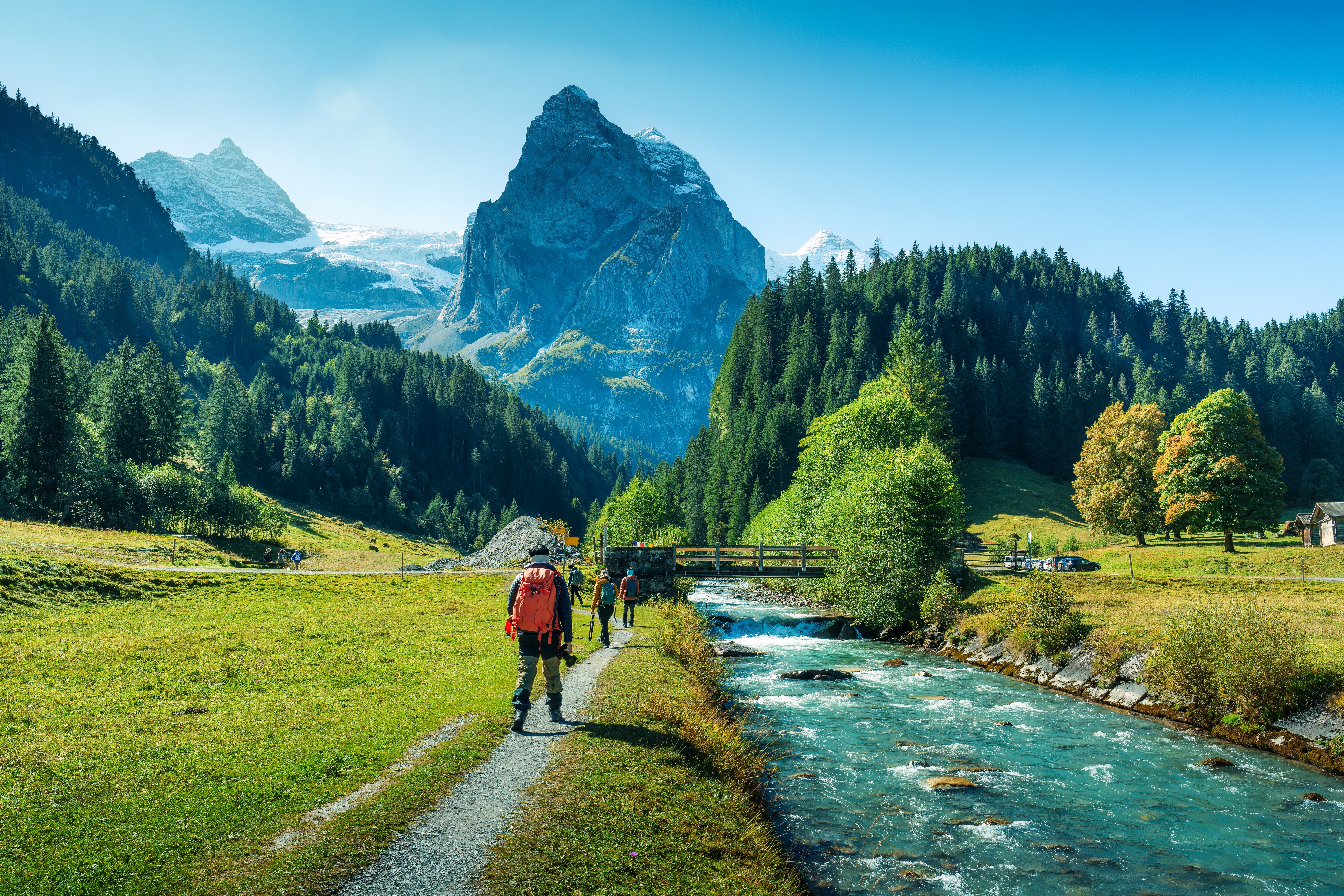 Vandrere nyder den smukke natur i Rosenlaui-dalen i Berner Oberland, Schweiz, med turkisblå bjergflod, sneklædte tinder og frodige grønne enge på en solrig sommerdag