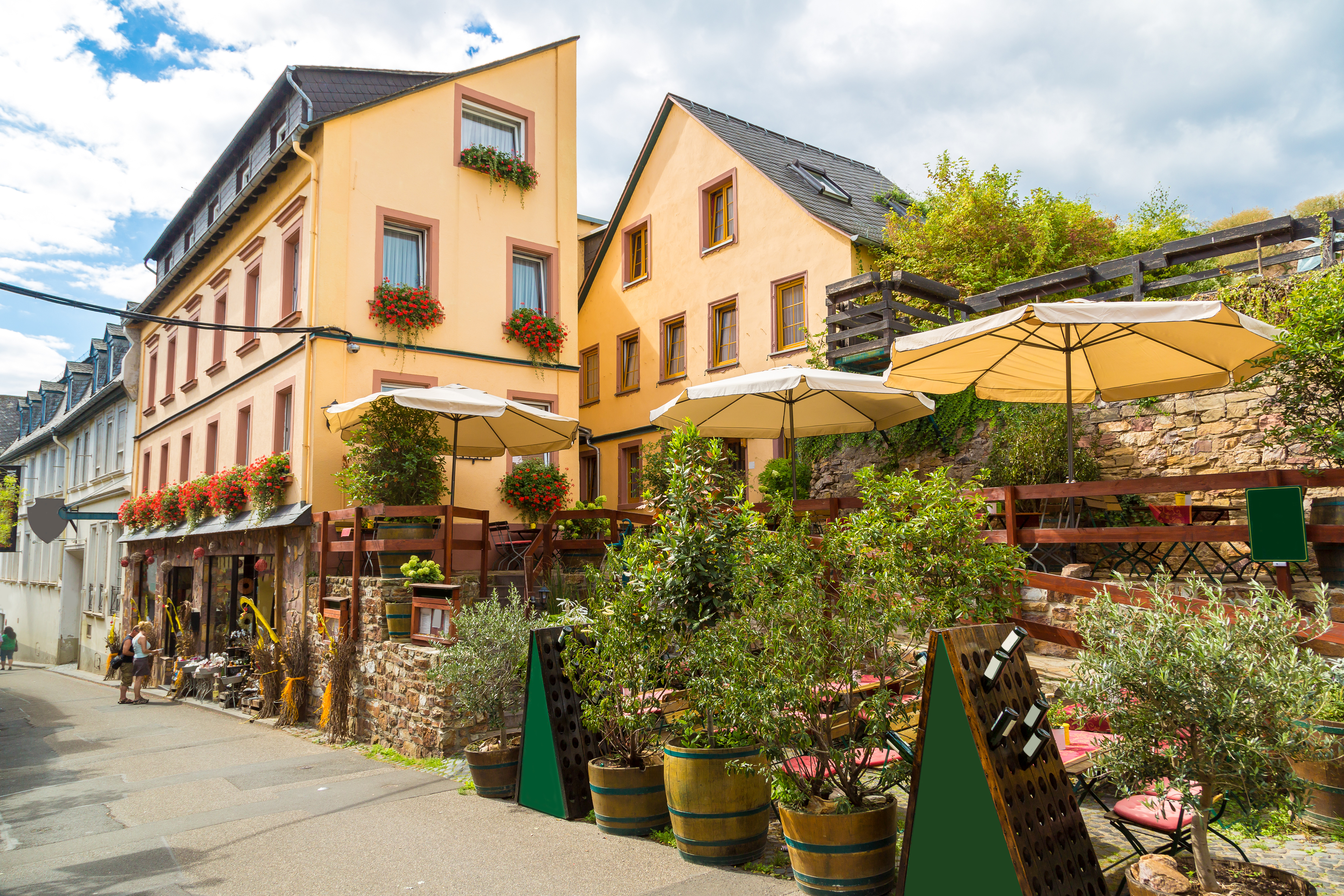 Idyllisk gadebillede fra Rüdesheim am Rhein med traditionelle tyske bygninger, blomstrende krukker og hyggelige udendørs caféer under gule parasoller i det berømte vinområde