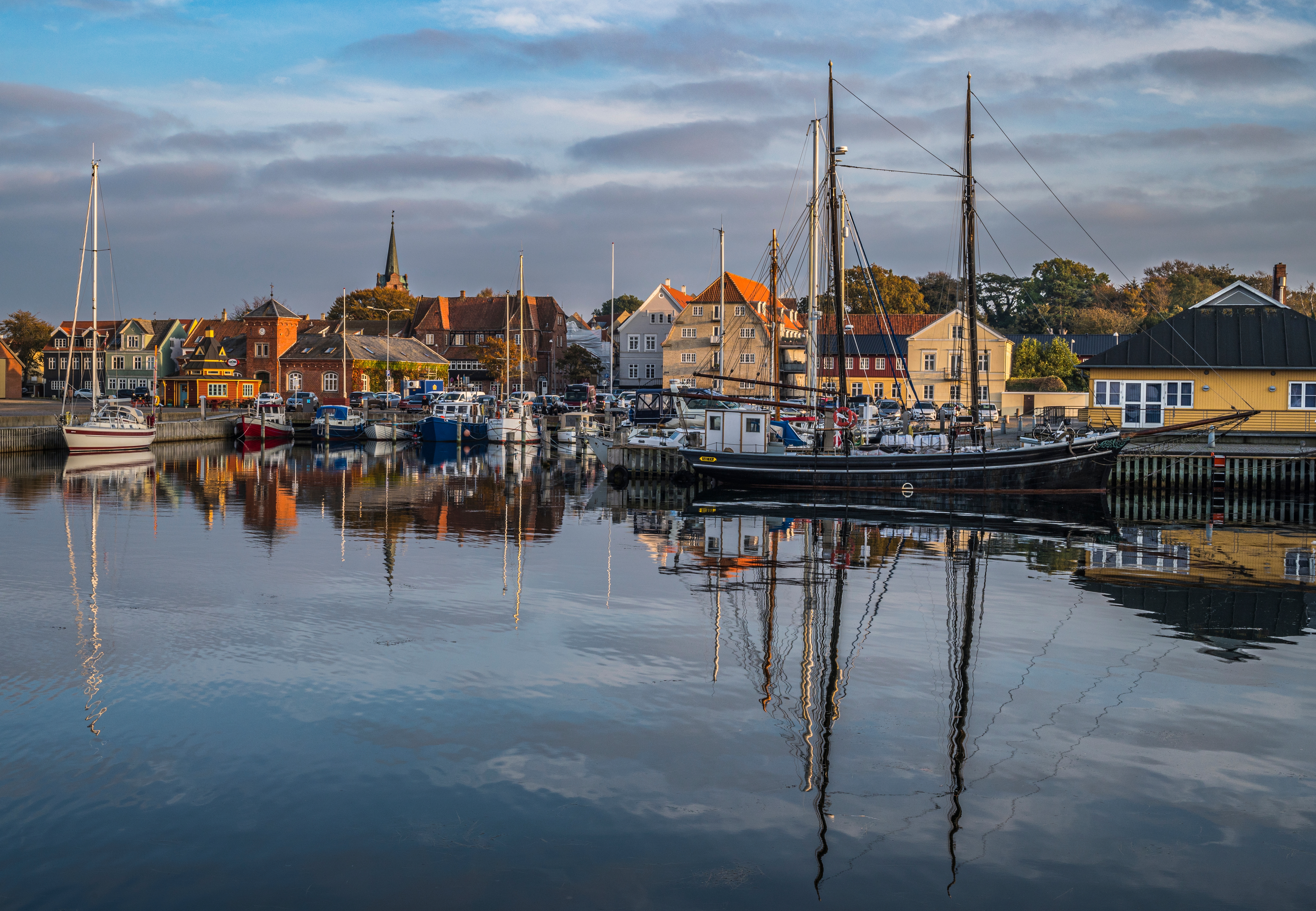 Smukt havnebillede fra Rudkøbing på Langeland med sejlbåde og farverige huse der afspejler sig i det blanke havnevand i solnedgangen