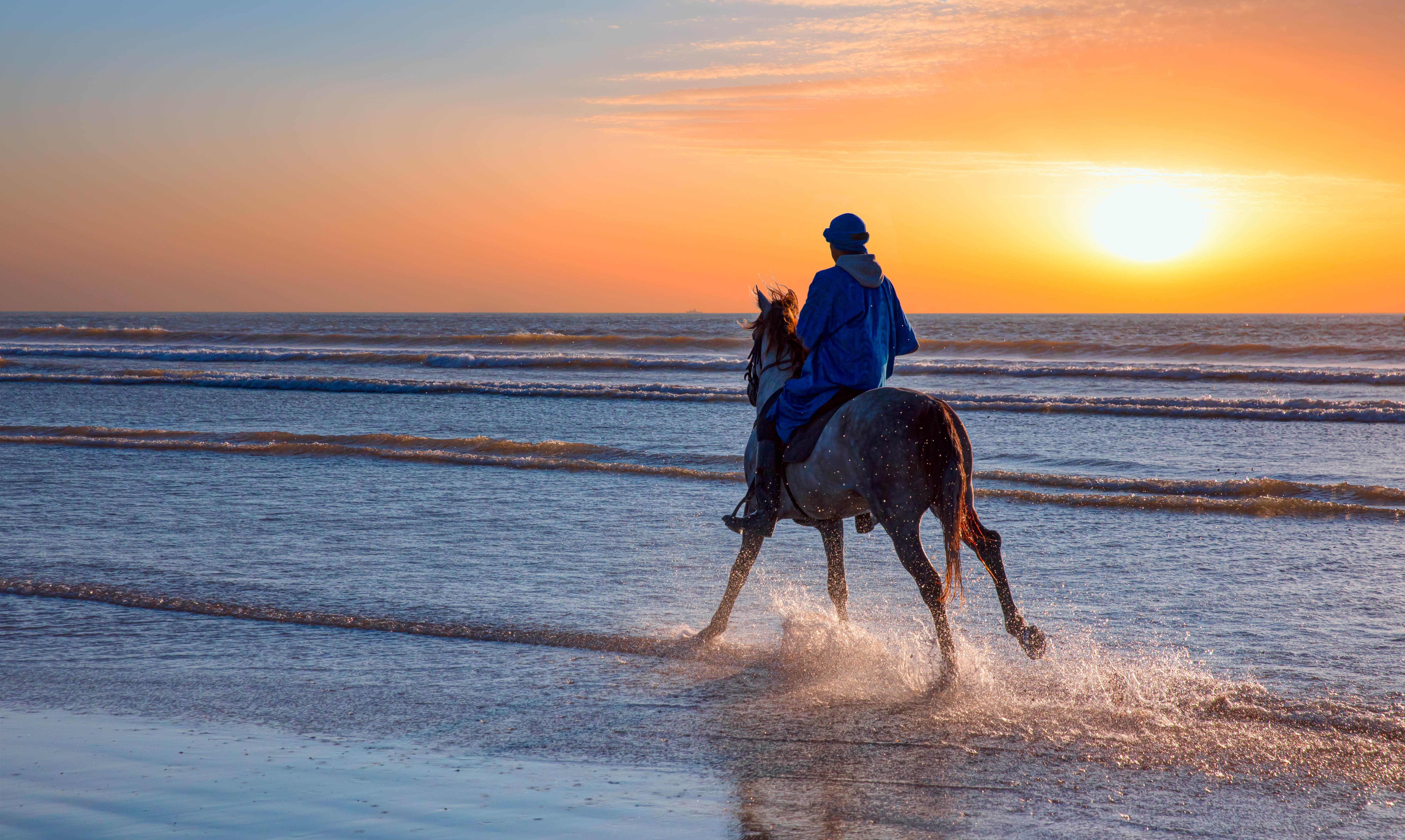 Hest og rytter på strand med sprøjtende havvand ved sunset