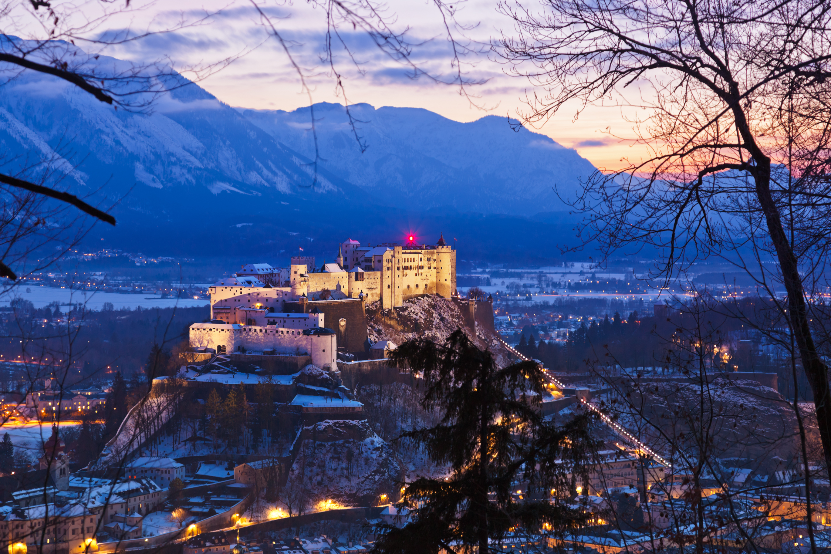 Smukt oplyst Hohensalzburg Fæstning i Salzburg ved solnedgang om vinteren med snedækkede Alper og byens glitrende lys