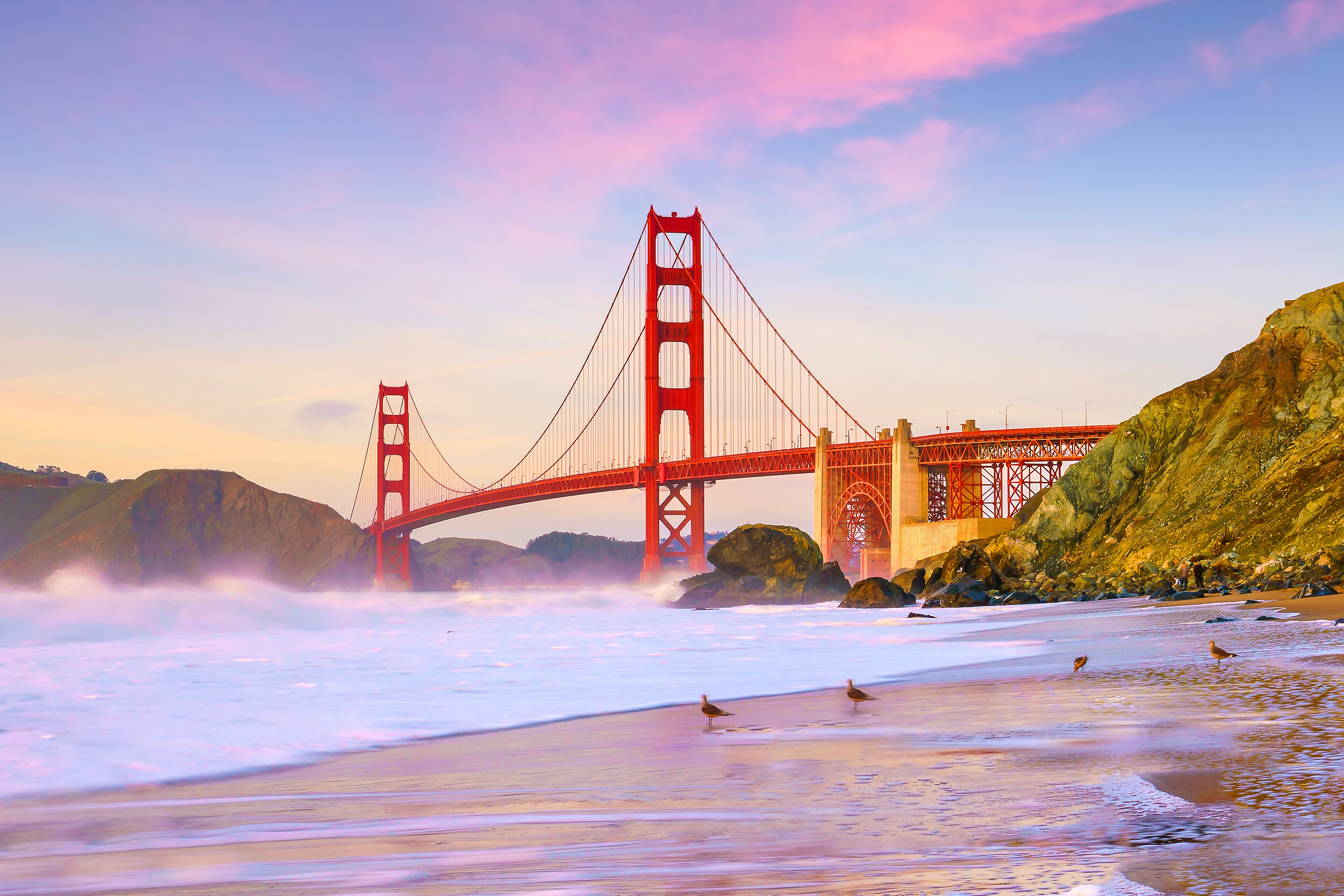Ikonisk Golden Gate Bridge i San Francisco set fra kysten ved solnedgang med dramatisk farverig himmel og bølger på stranden