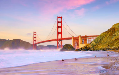 Ikonisk Golden Gate Bridge i San Francisco set fra kysten ved solnedgang med dramatisk farverig himmel og bølger på stranden