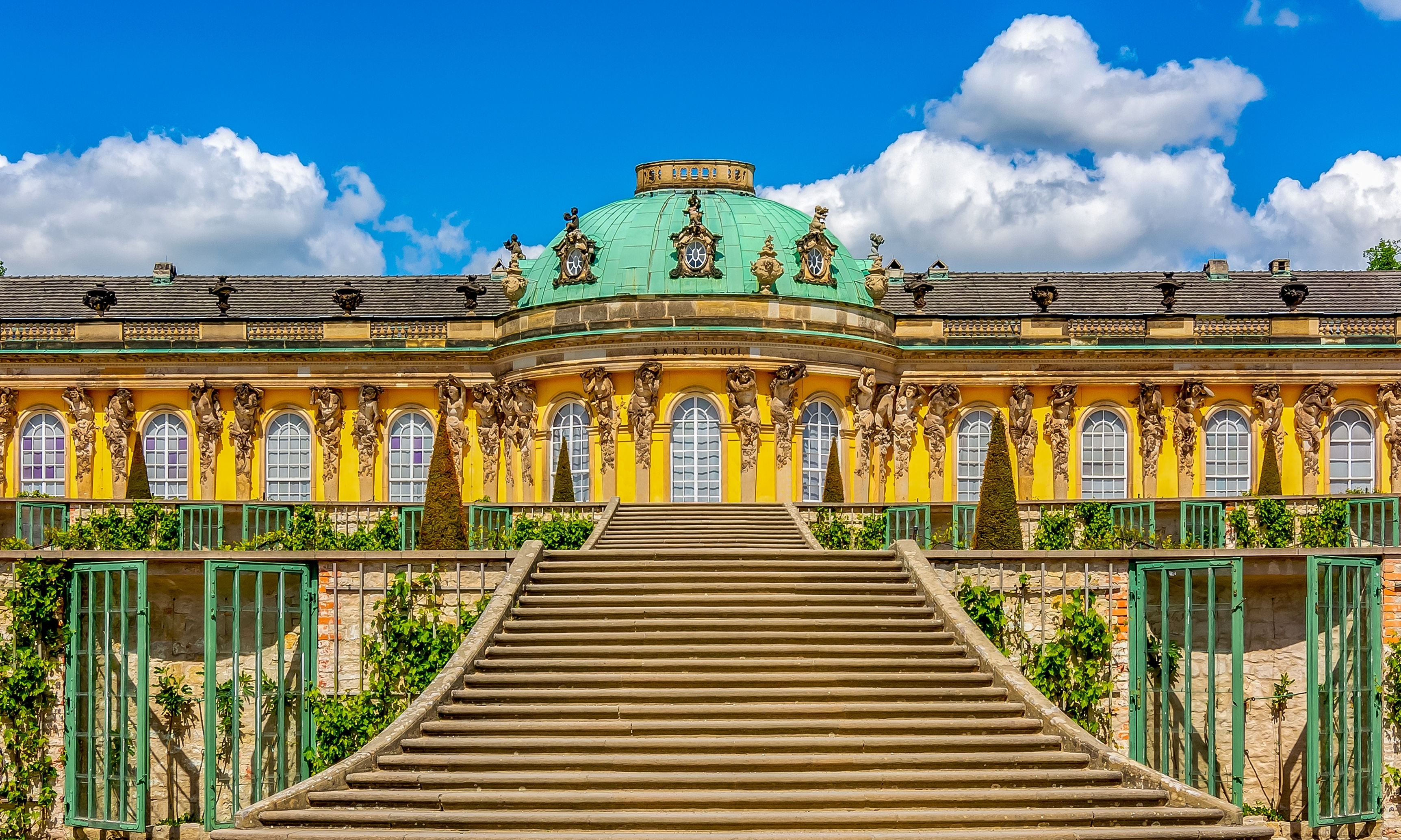 Sanssouci Slot i Potsdam om sommeren – barok arkitektur med gul facade, grøn kuppel og bred trappe op til det historiske palads