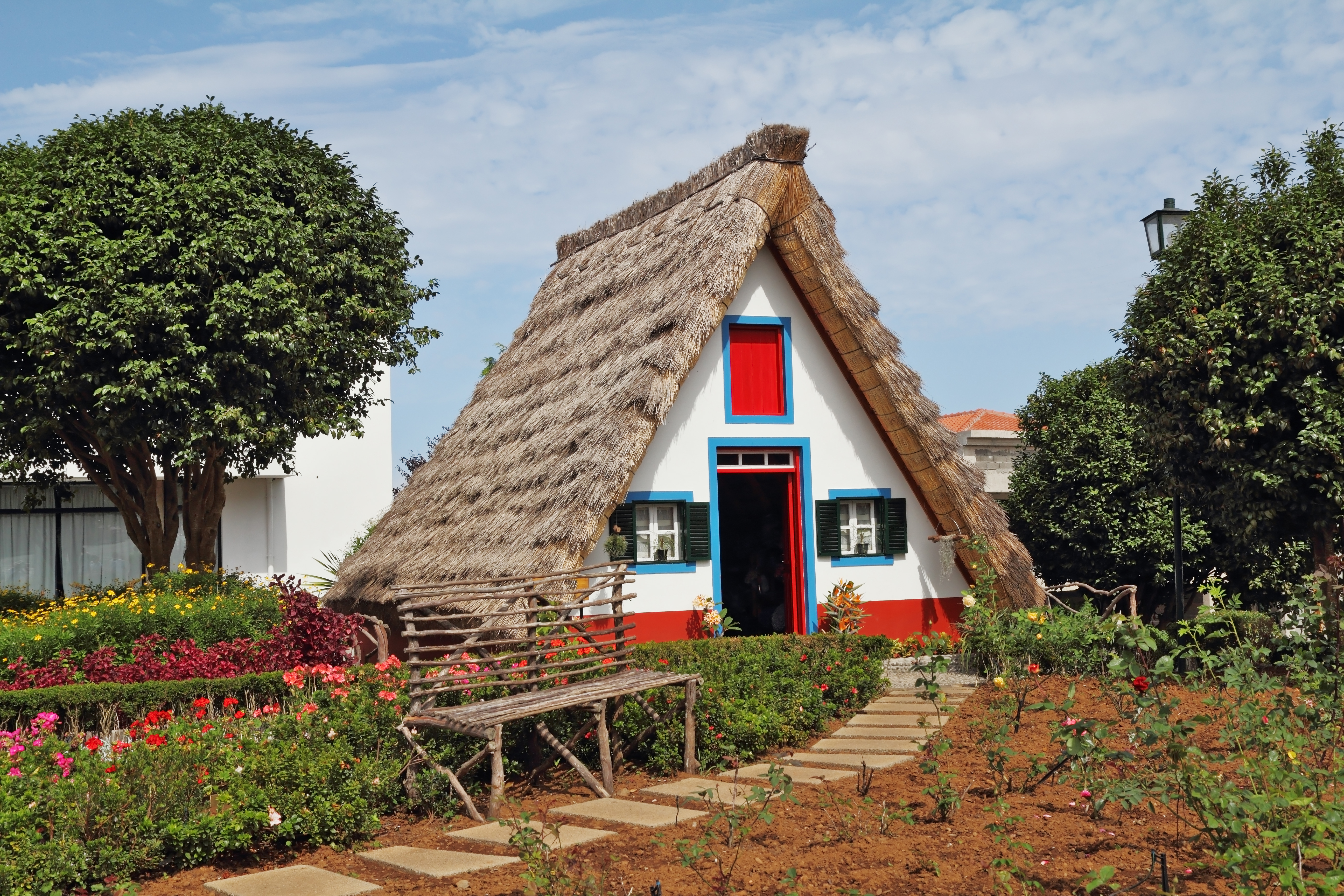 Idyllisk triangulært hus med stråtag og iøjnefaldende rød dør i Santana på Madeira, omgivet af frodige blomster og traditionel portugisisk landsbystemning