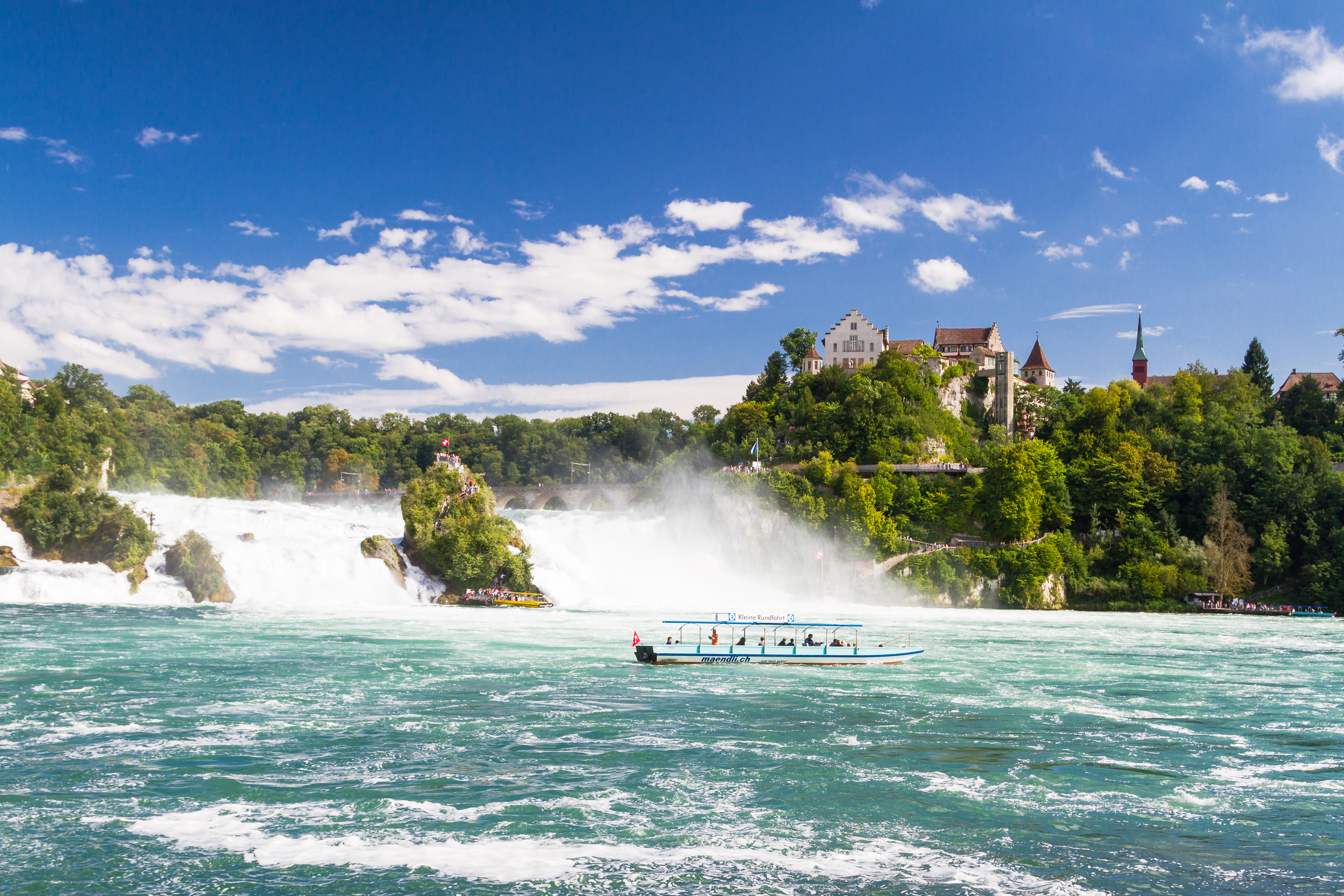 Storslået udsigt over Europas største vandfald, Rhinfaldet i Schaffhausen, Schweiz, med dets dramatiske vandmasser der falder over klippeformationer, skaber hvidt skum og spraytåge, mens en turistbåd giver perspektiv til den imponerende naturkraft