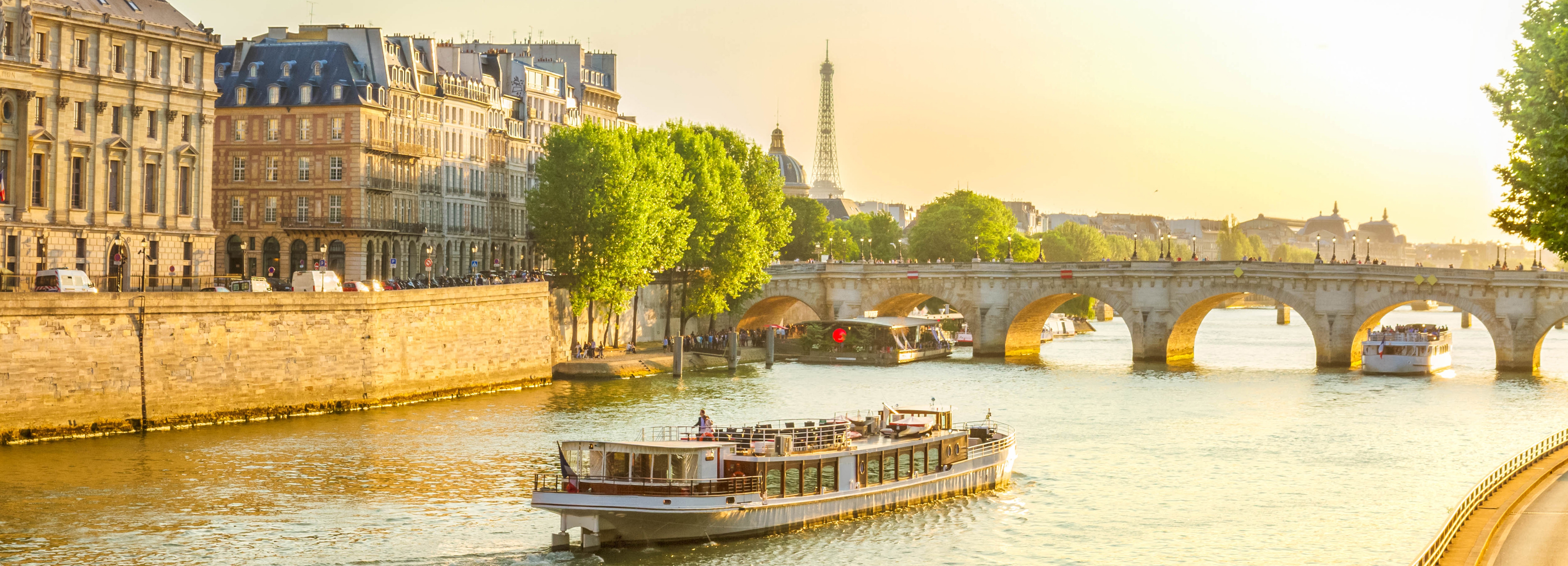 Smukt udsyn over Pont Neuf og Seinen i Paris med krydstogtbåd og gyldent solnedgangslys samt Eiffeltårnet