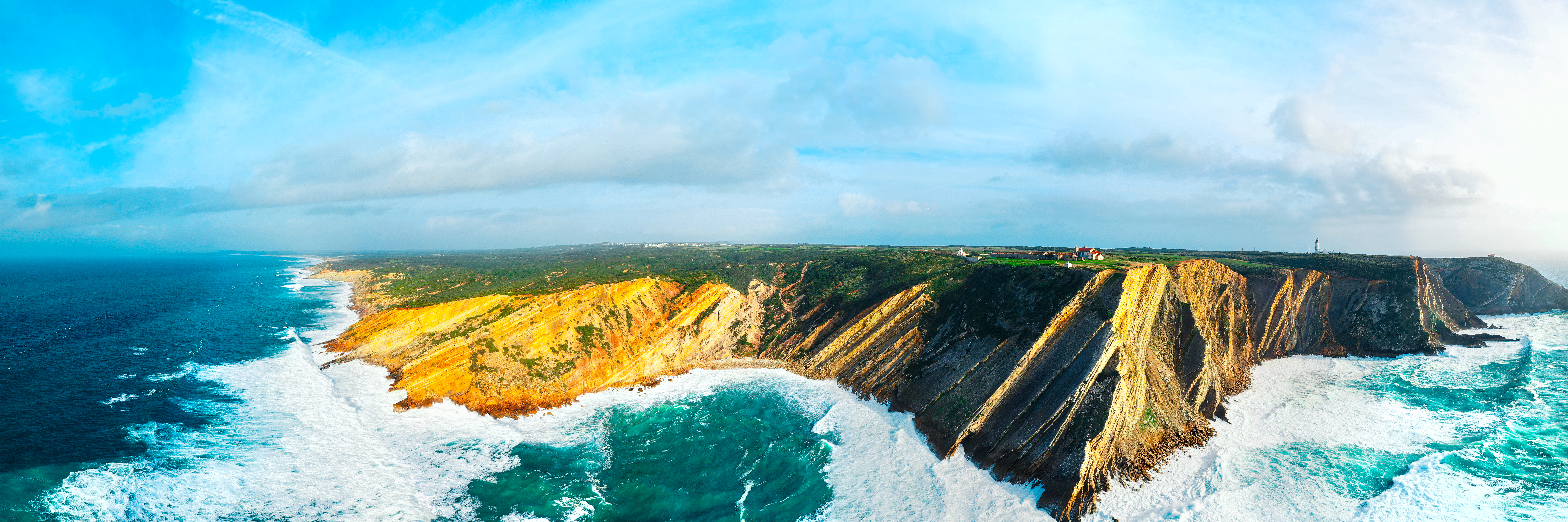Flot fugleperspektiv over Cabo Espichel ved Sesimbra i Portugal med dramatiske kystklinter, kraftige atlanterhavsbølger og grønt klippelandskab under blå himmel