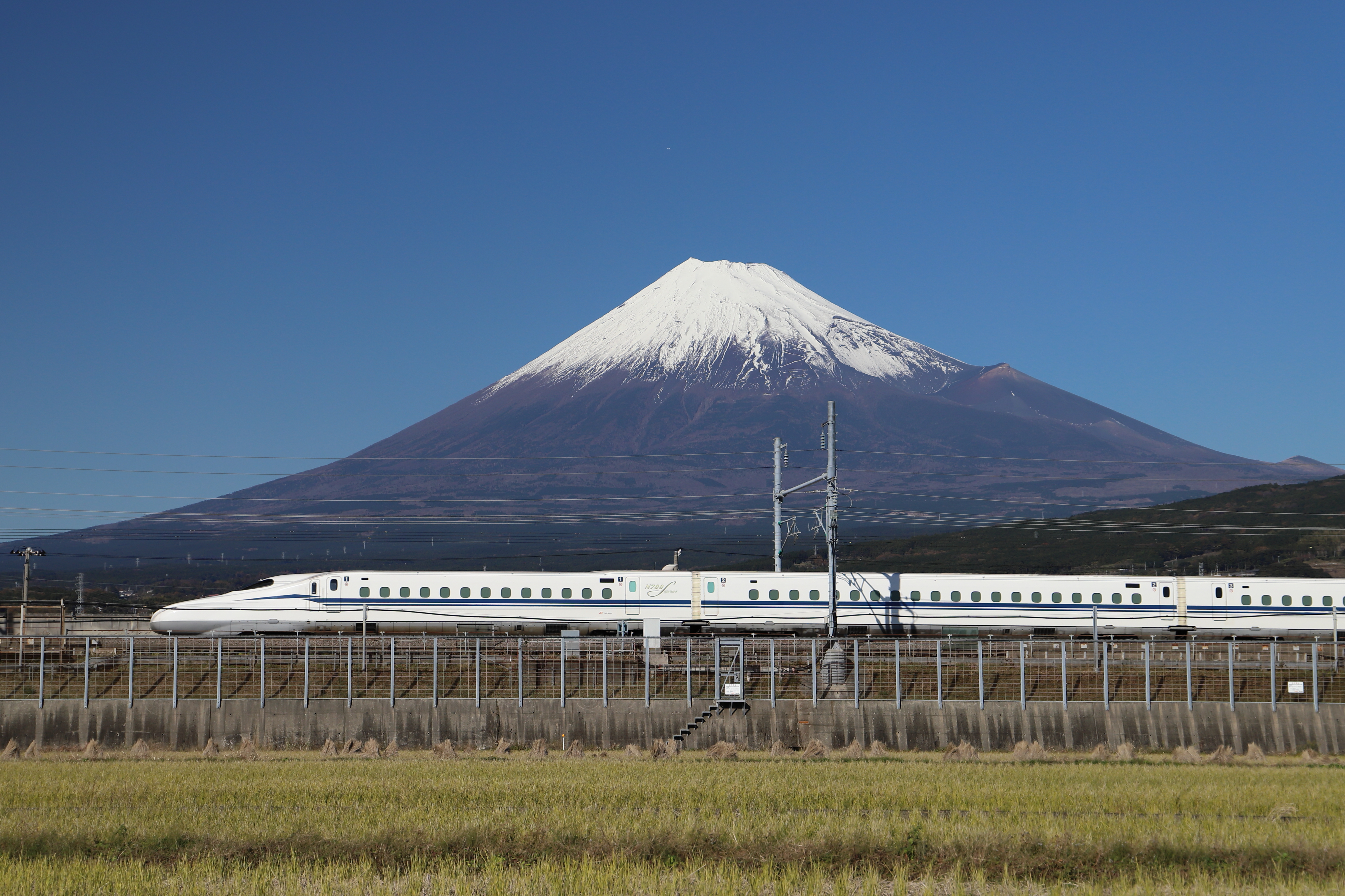 Fascinerende syn af Japans højteknologiske Shinkansen-hurtigtog, der kører gennem landskabet med det snedækkede Fuji-bjerg, der majestætisk tårner sig op i baggrunden mod en perfekt blå himmel