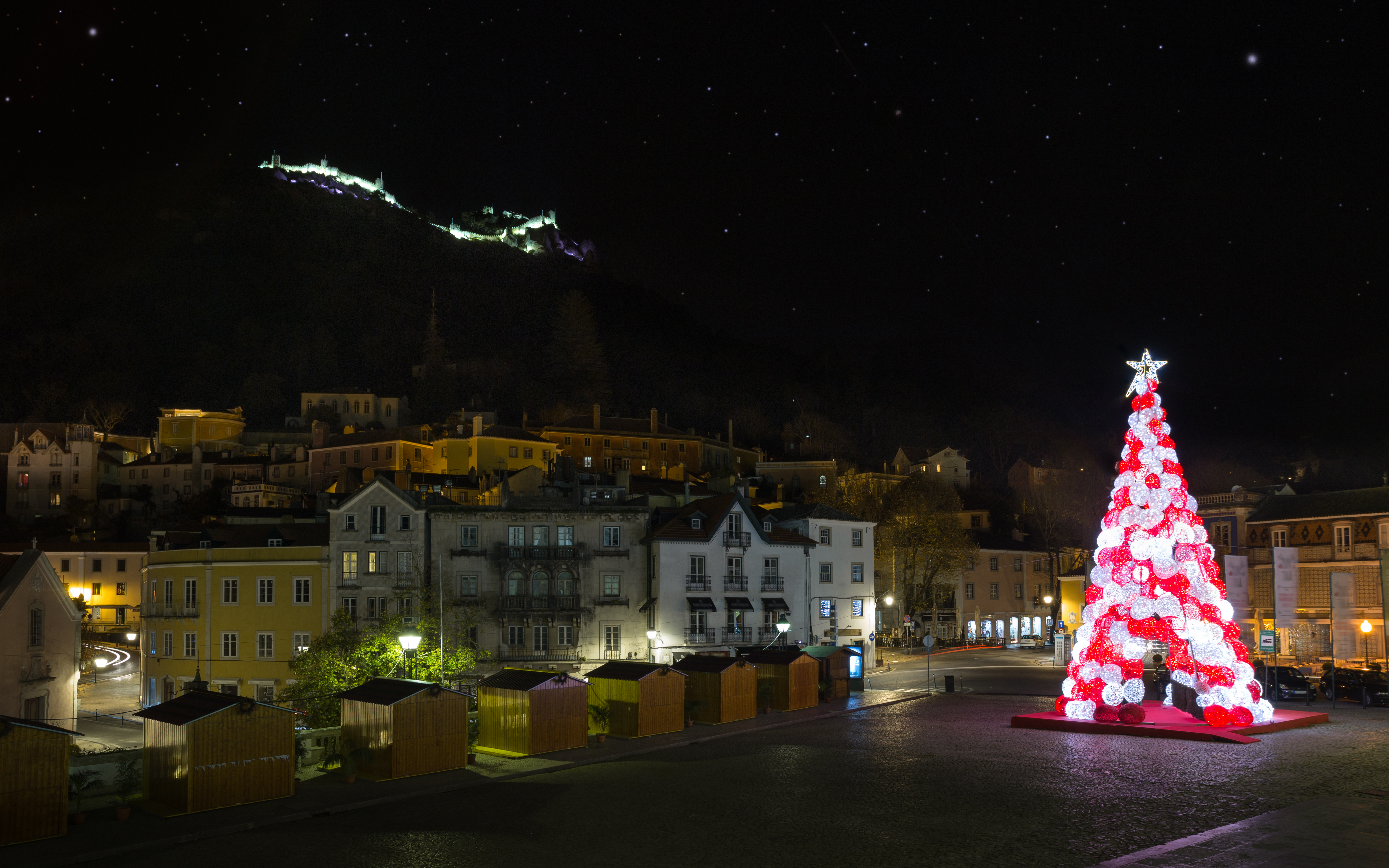 Smukt oplyst juletræ med røde og hvide kugler på torvet i Sintra med det historiske slot og stjerneklar himmel om natten i Portugal