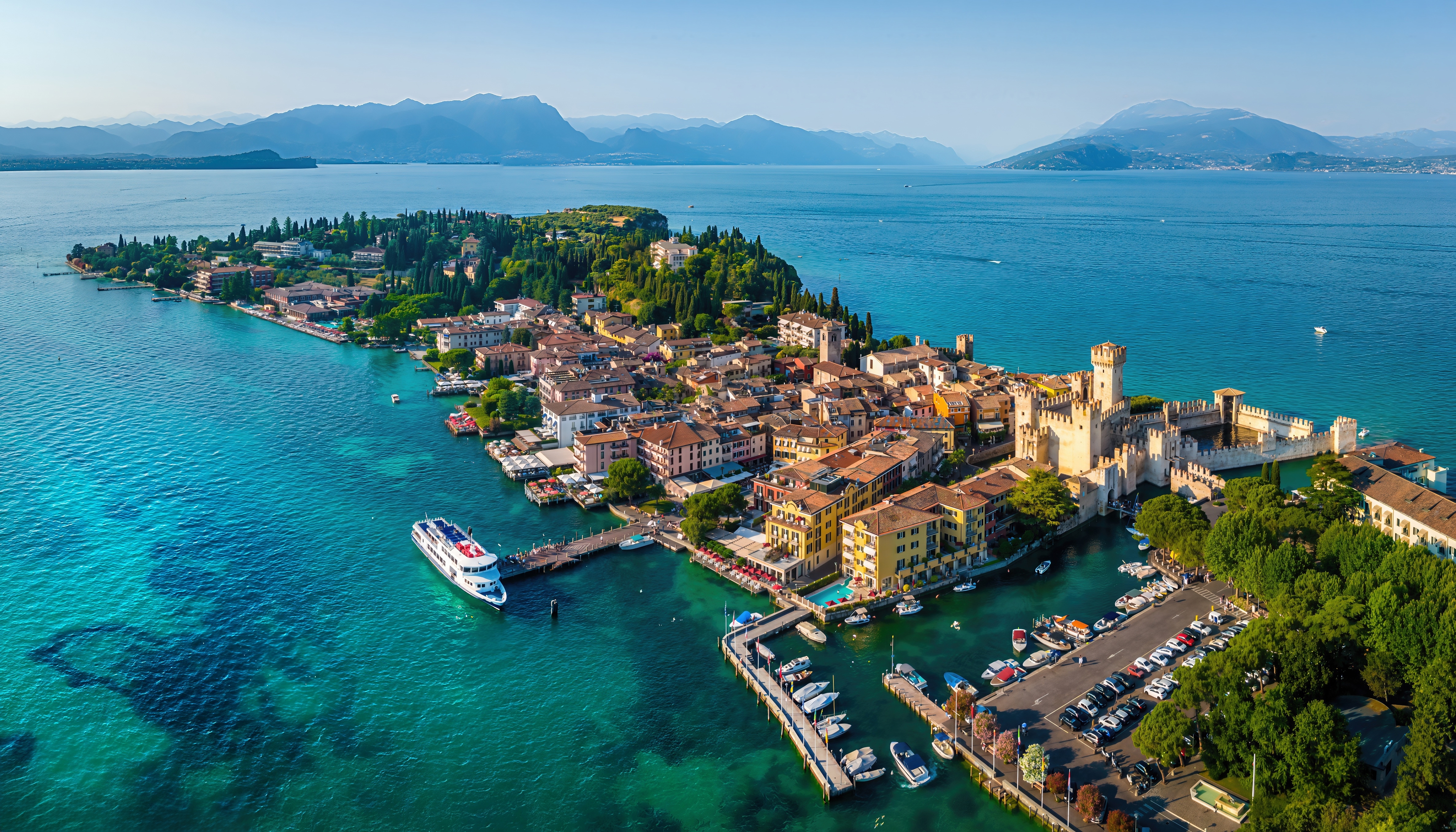 Dronefotos af den maleriske Sirmione-halvø ved Gardasøen med det ikoniske Scaliger-slot, hyggelig havn og azurblåt vand mod en bjergrig horisont